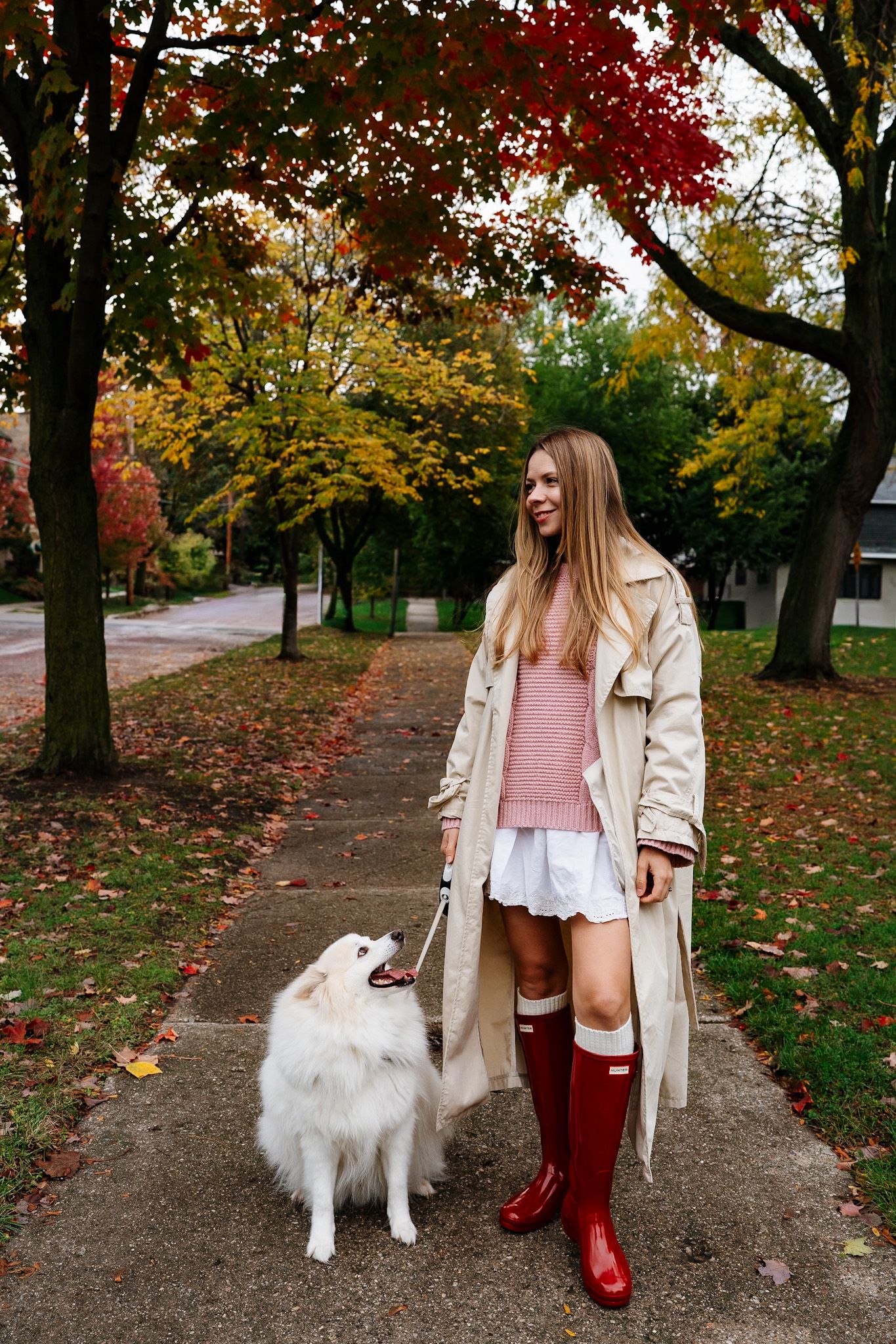 Woman in trench coat and red boots walks white fluffy dog on autumn path with colorful fall foliage.