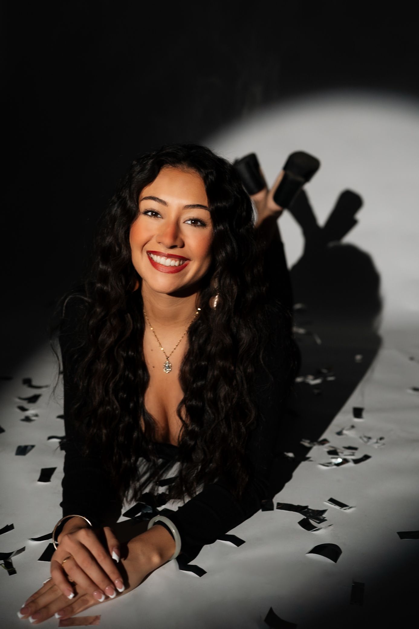 Studio portrait with long dark hair and jewelry against dramatic lighting and confetti backdrop.