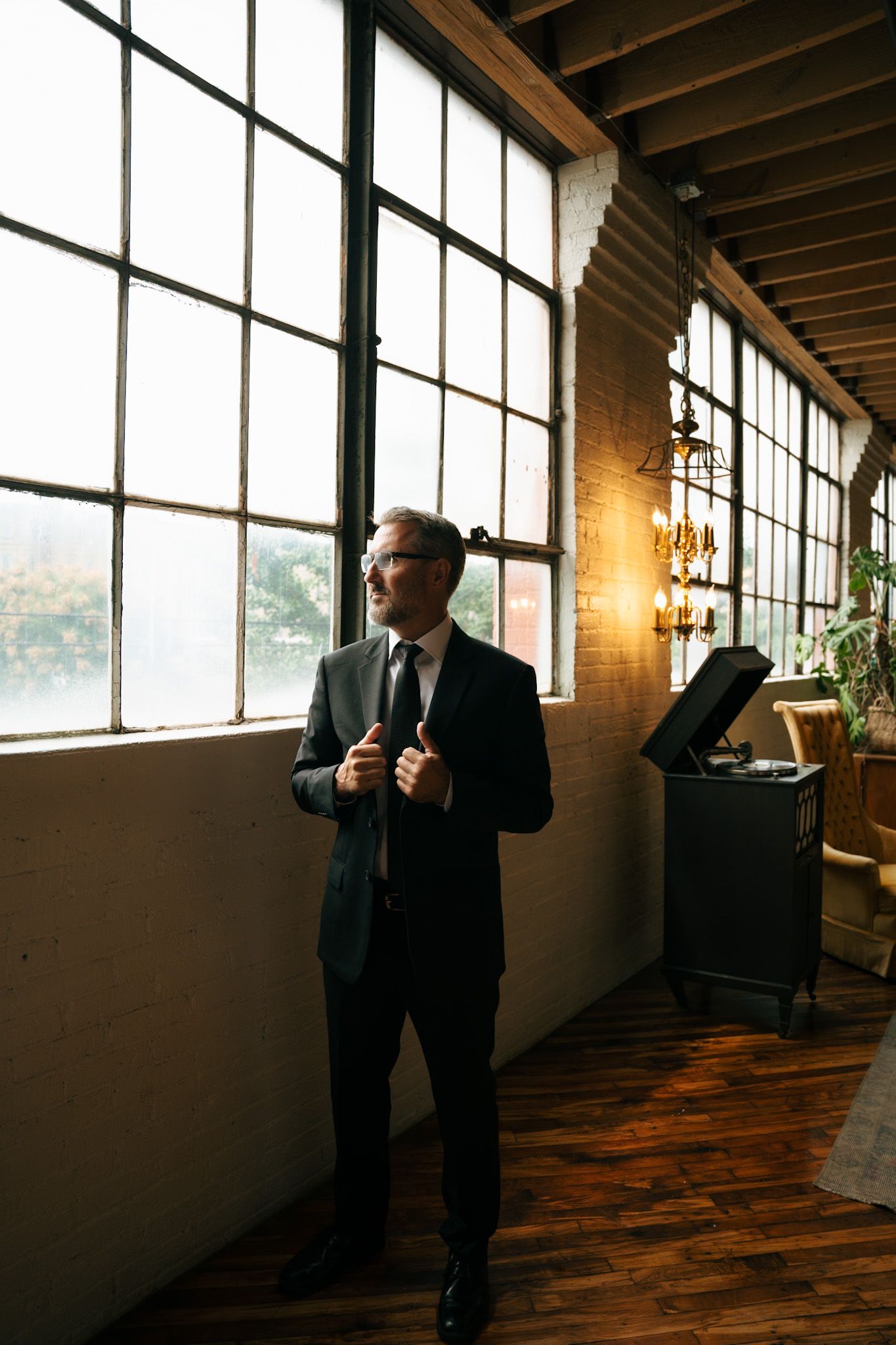 Professional in a black suit poses against industrial windows and exposed brick in a loft-style space.