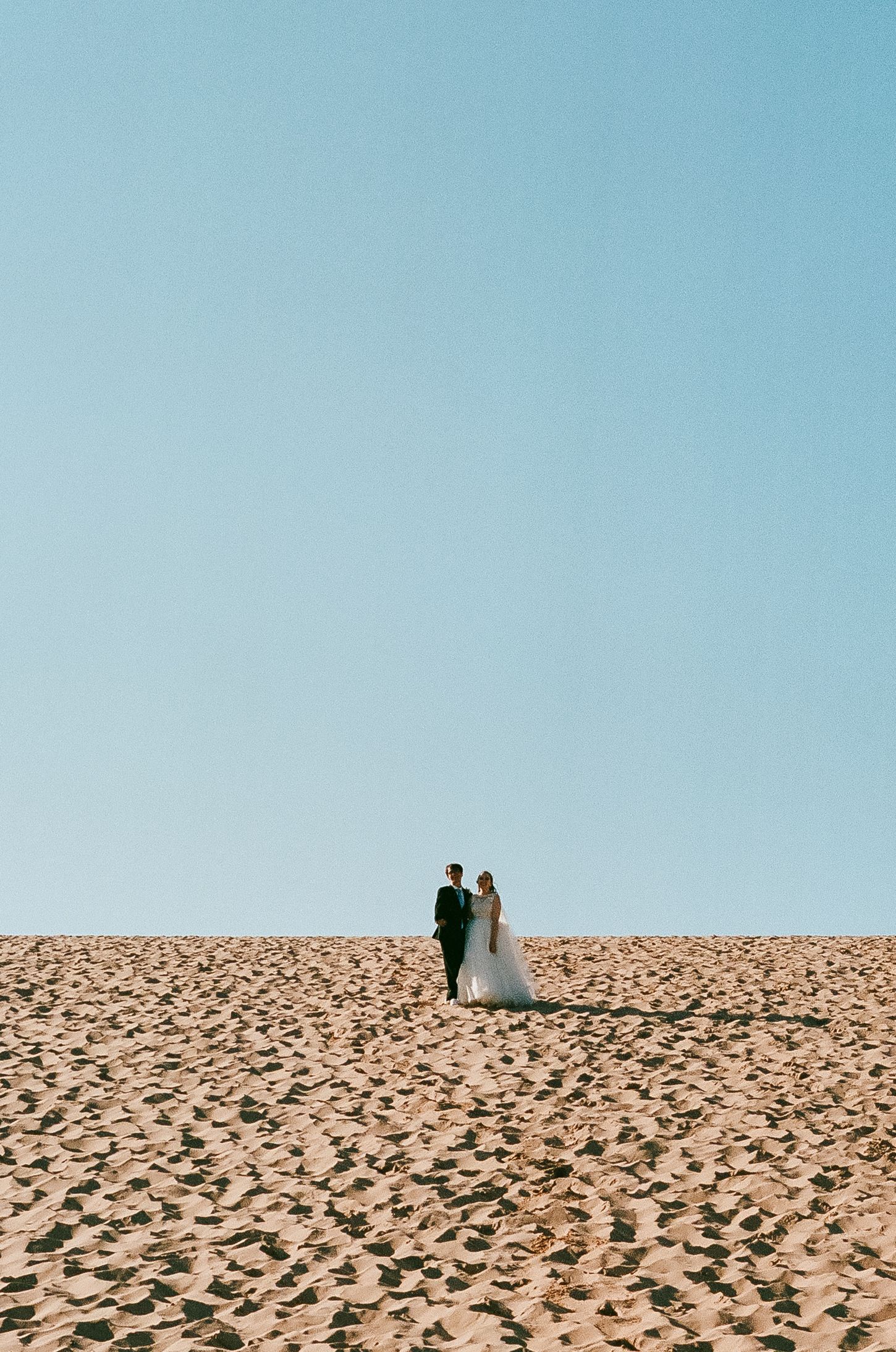 Couple walks along sandy dune against clear blue sky.