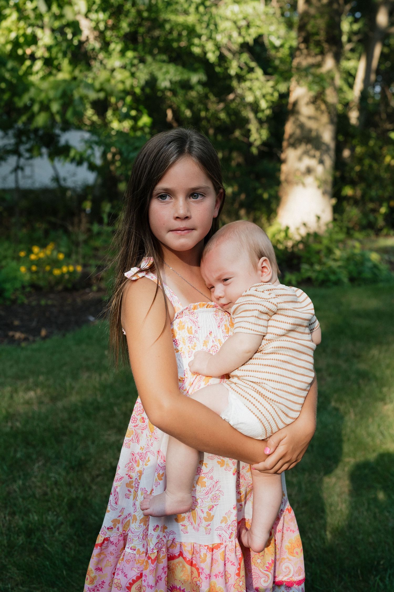 A young girl holds a sleeping baby in a floral dress against a green garden background.