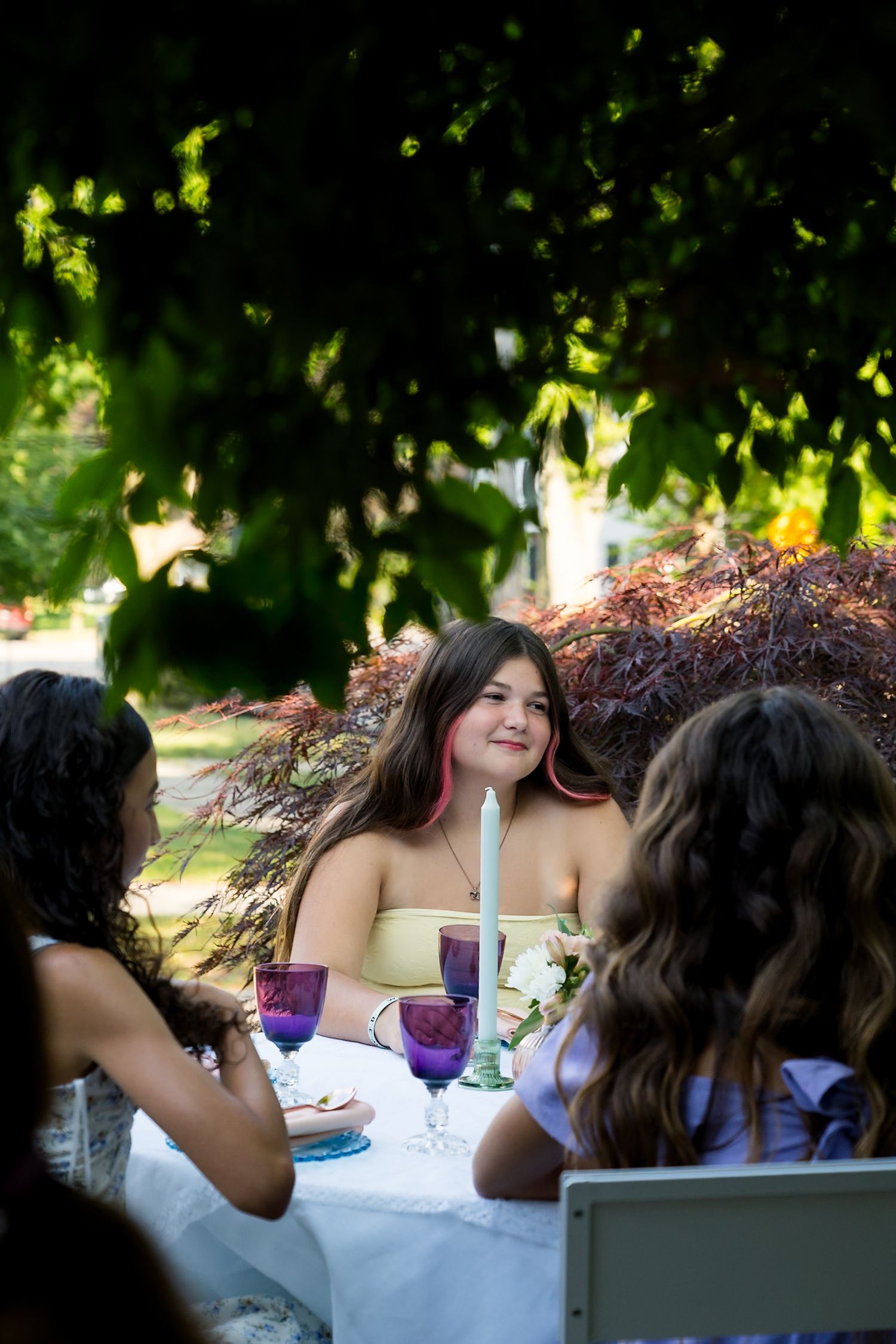 Friends gathered around outdoor table under dappled sunlight and trees.