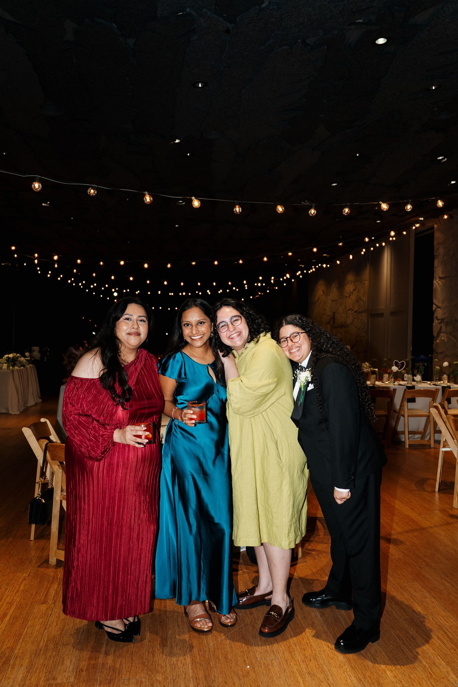 A group of wedding guests in formal attire posing under string lights at a reception.