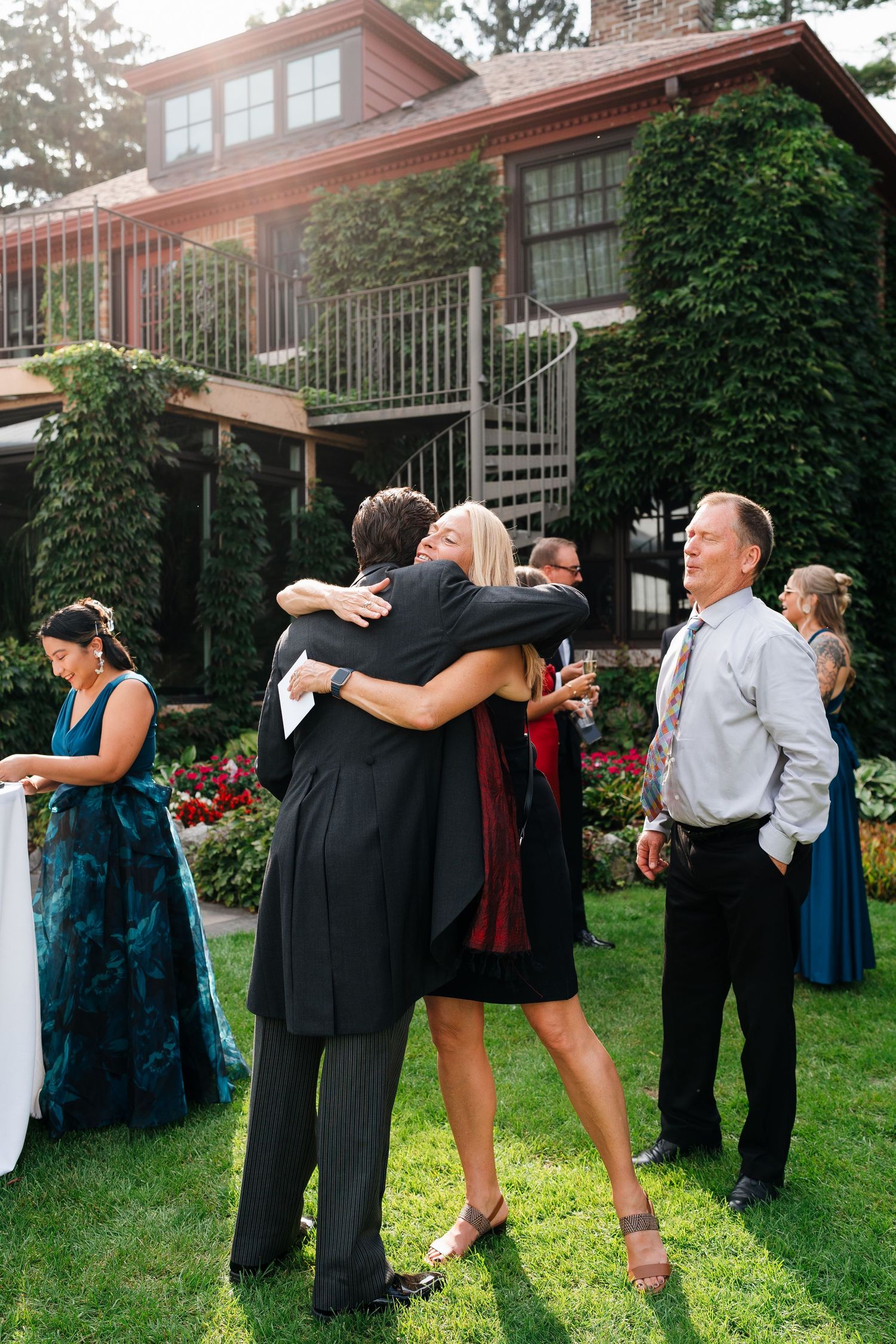 Wedding guests embracing and socializing on a lawn outside a building during a celebration.