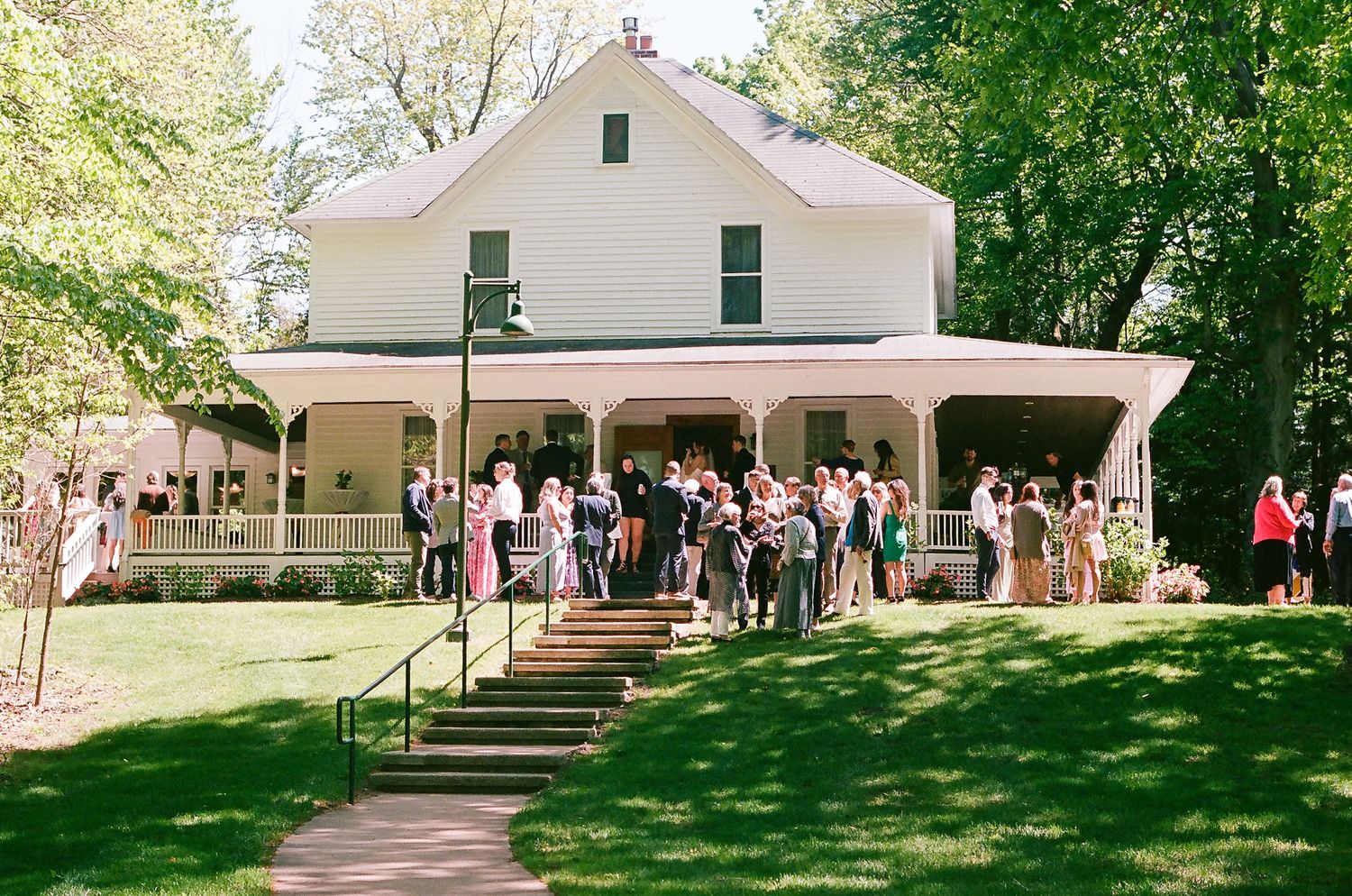 A charming white farmhouse with a wraparound porch hosts wedding guests gathering on its front steps.