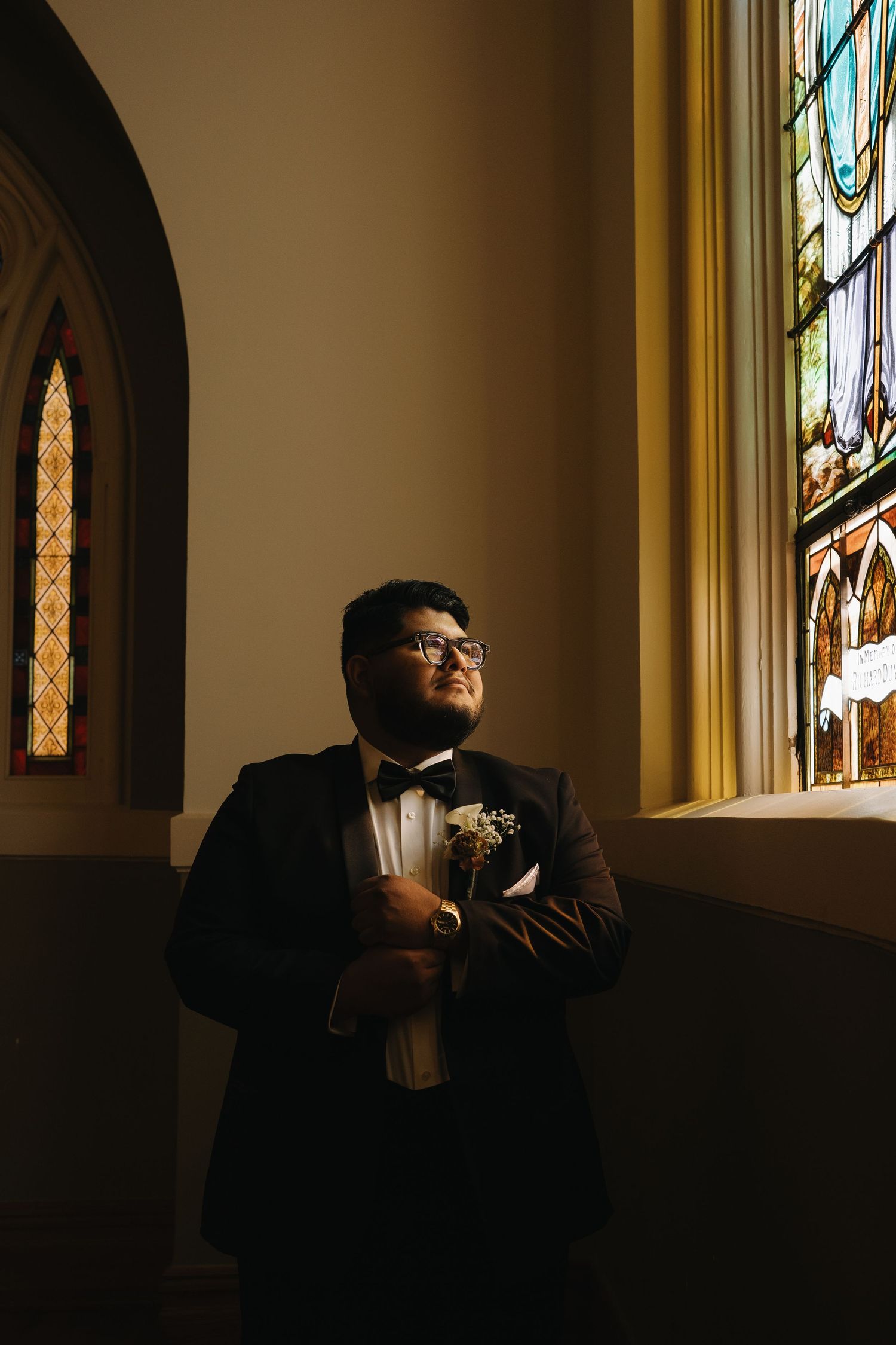 A person in formal attire stands in dramatic lighting near church windows.