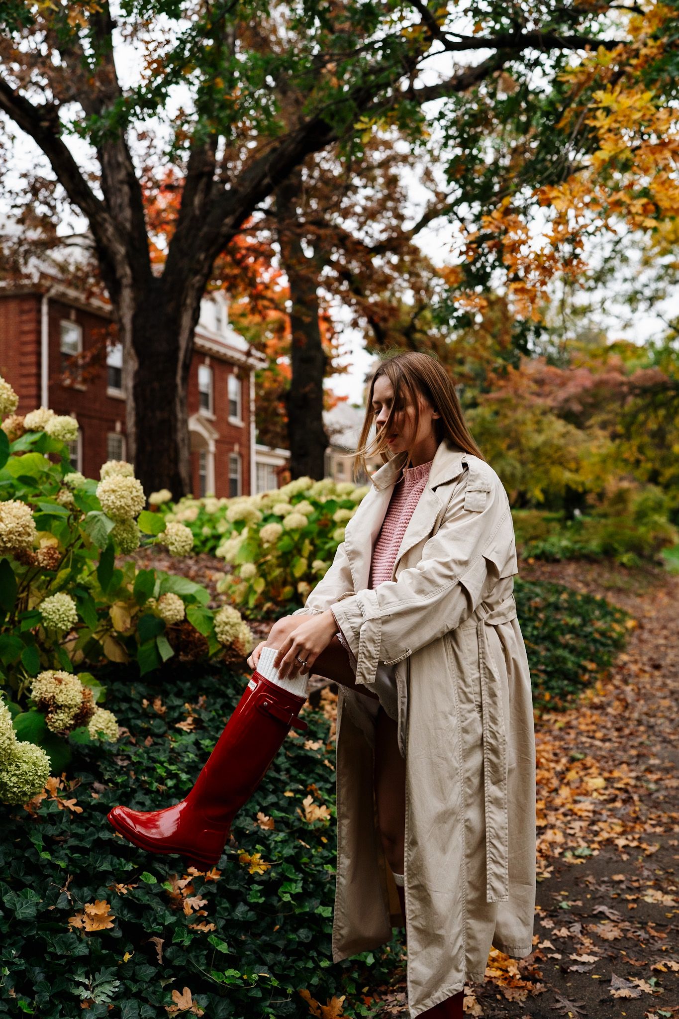 Red boots and beige trench coat styled against autumn leaves and hydrangea bushes in a garden setting.