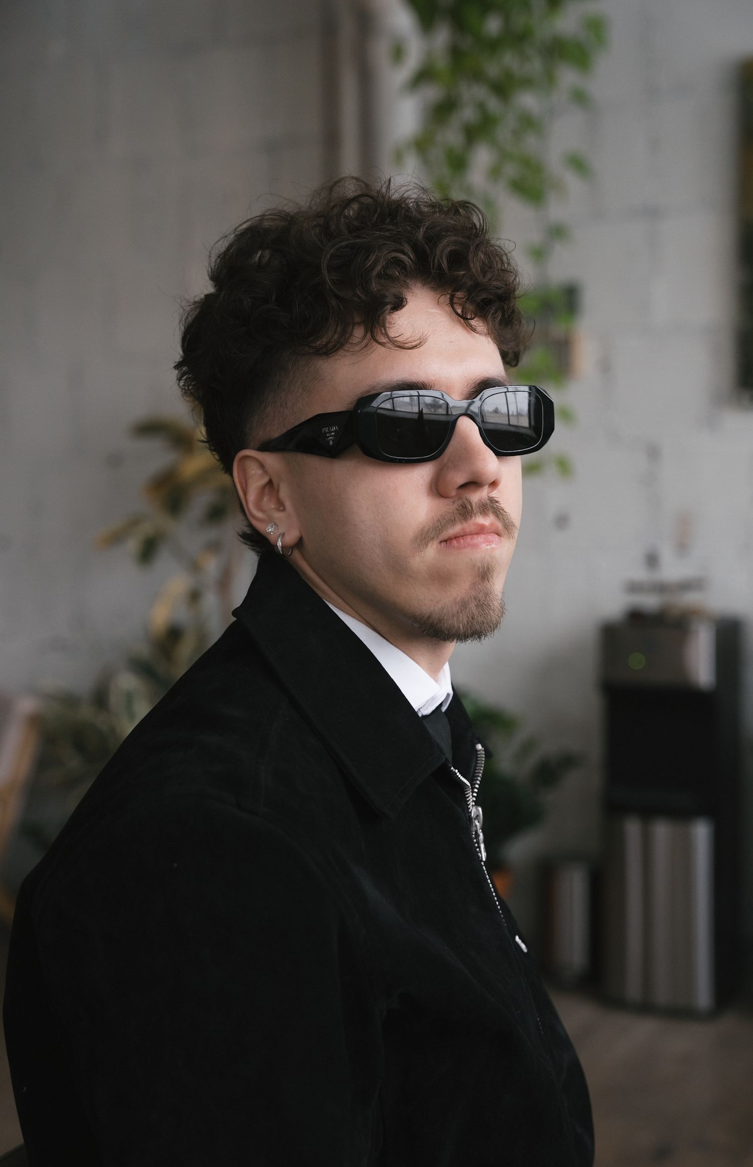 Fashion model wearing black coat and sunglasses poses against light background in artistic portrait series.