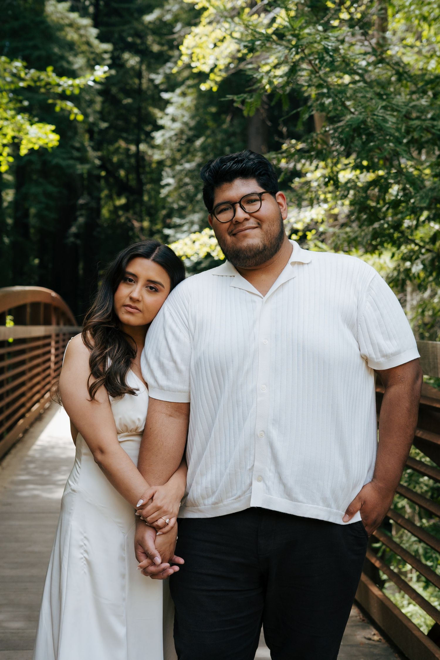 A couple holding hands while standing on a wooden bridge surrounded by lush forest greenery.