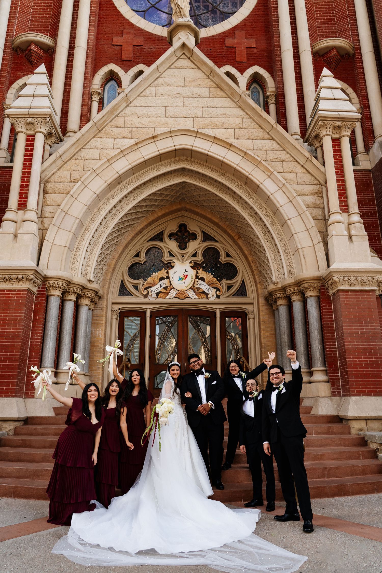 Wedding party gathered on stone steps of Gothic revival church with red brick and limestone details.