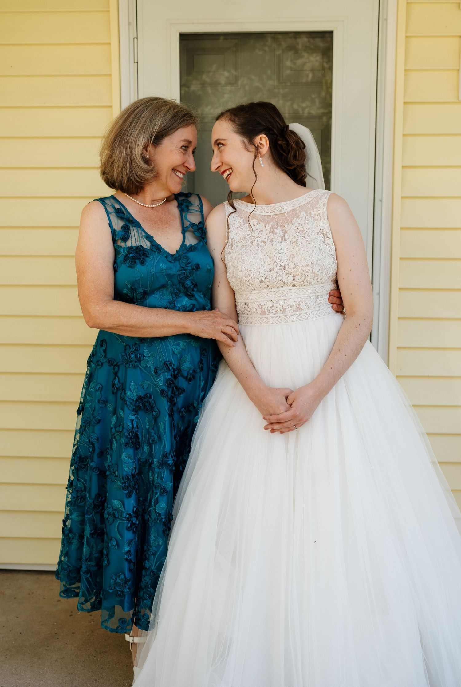 Bride in white gown shares tender moments with bridesmaid in teal dress against yellow house exterior.