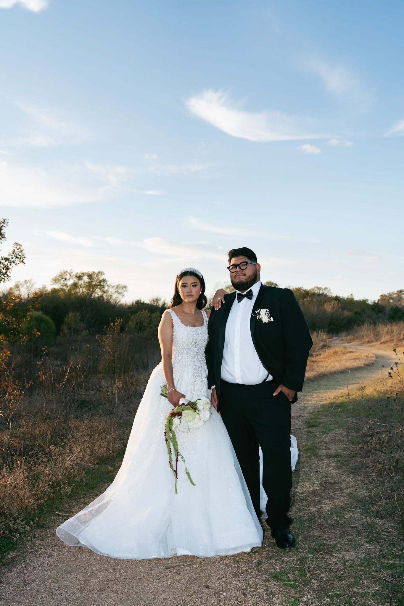 A couple in wedding attire poses together on a scenic hillside path during golden hour.