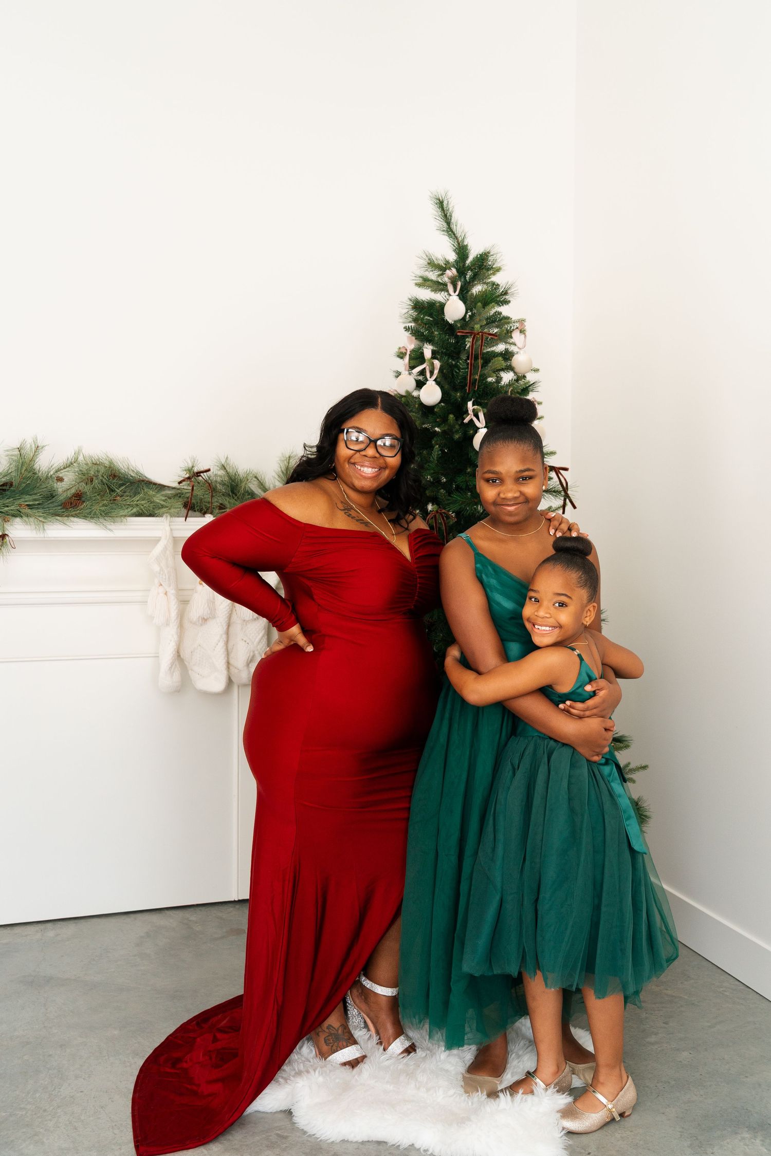 A group poses in formal dresses against a white wall with Christmas decorations.