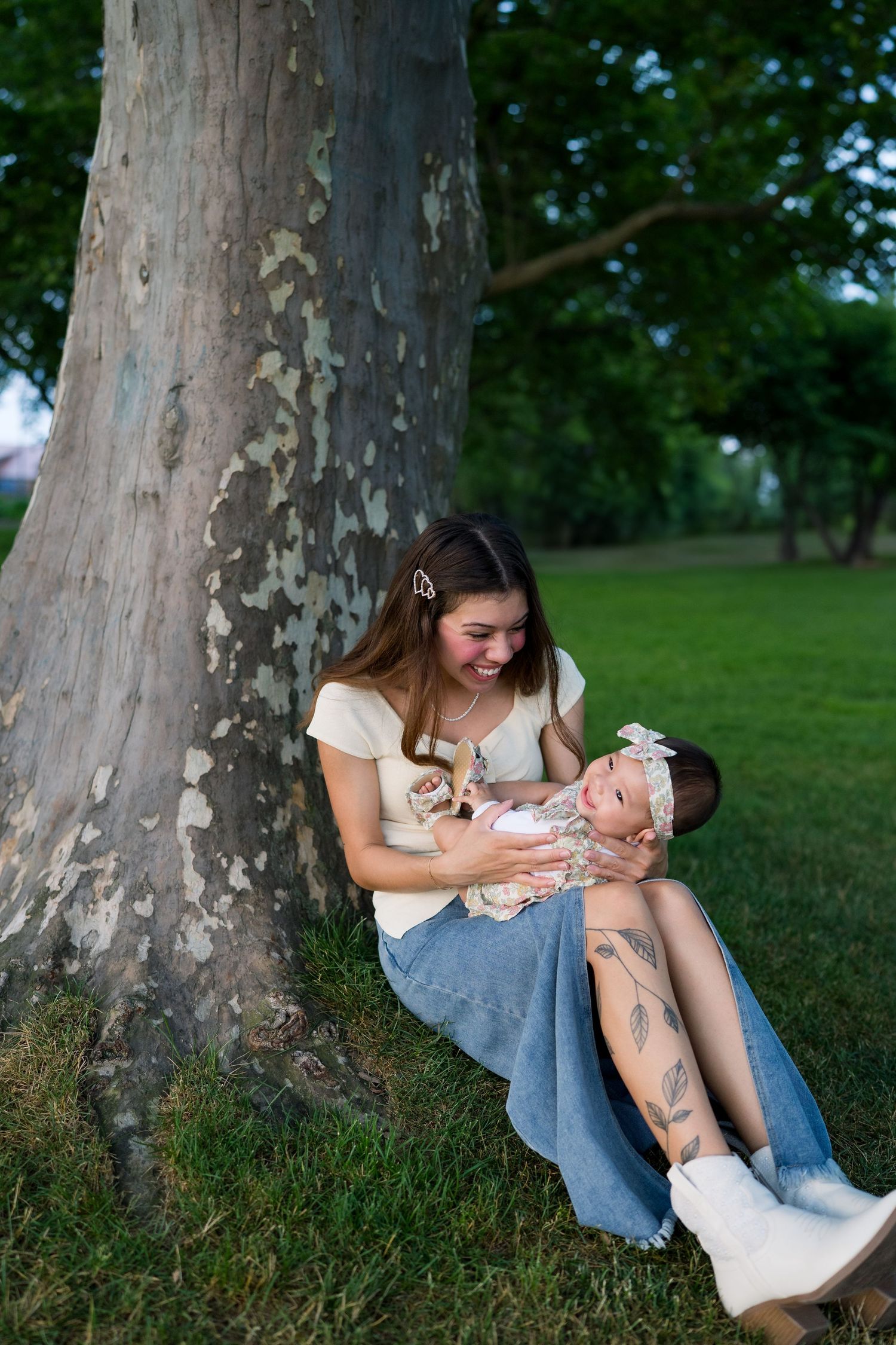 Mother and baby share tender moment while sitting under large tree in park.