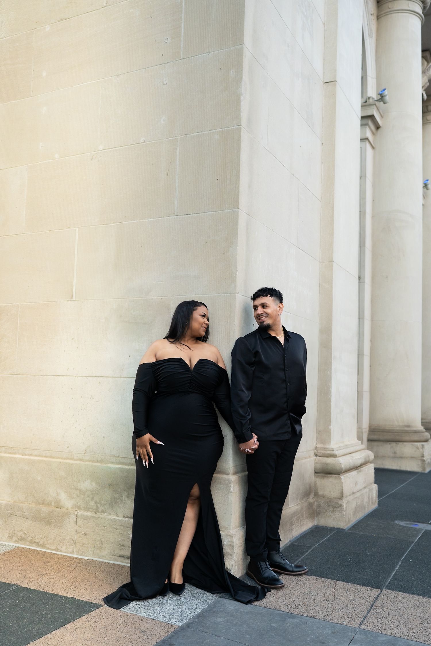 A couple in matching black outfits poses casually against white architectural columns.
