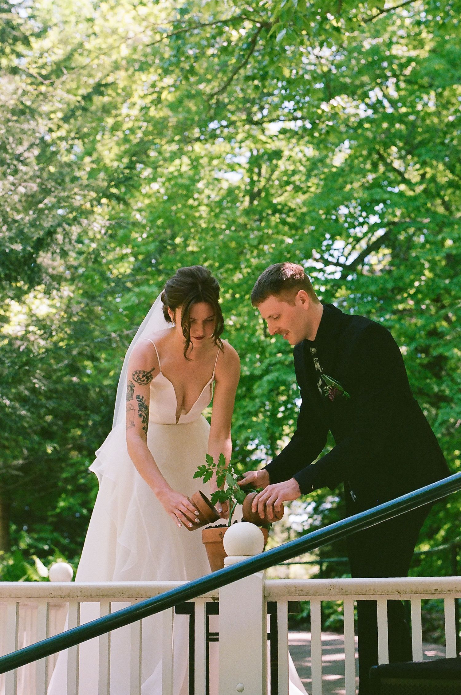 Couple leaning over a white railing outdoors surrounded by green trees.