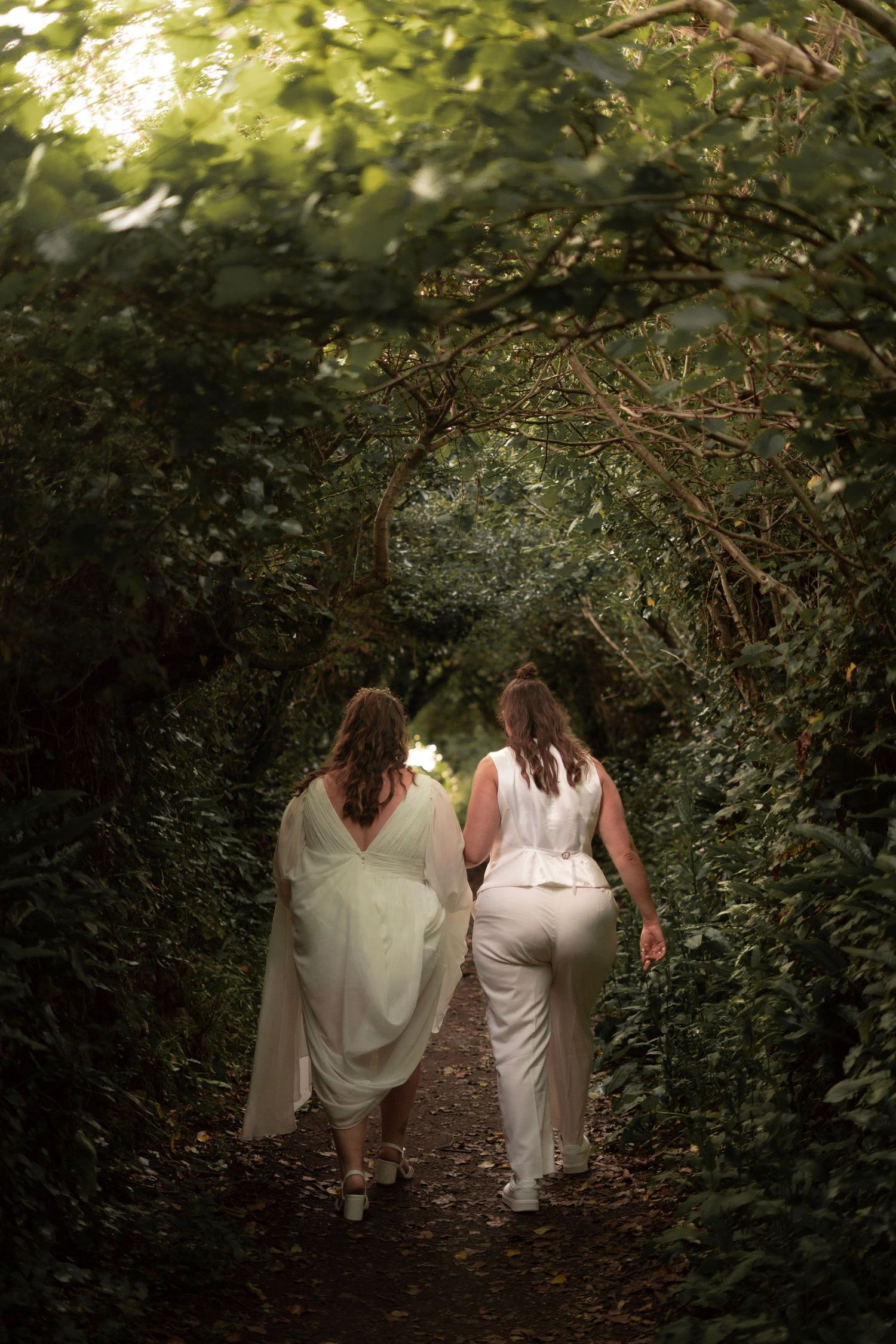 Two brides walking through the woodland arch at Lantic Bay near Tredudwell Manor