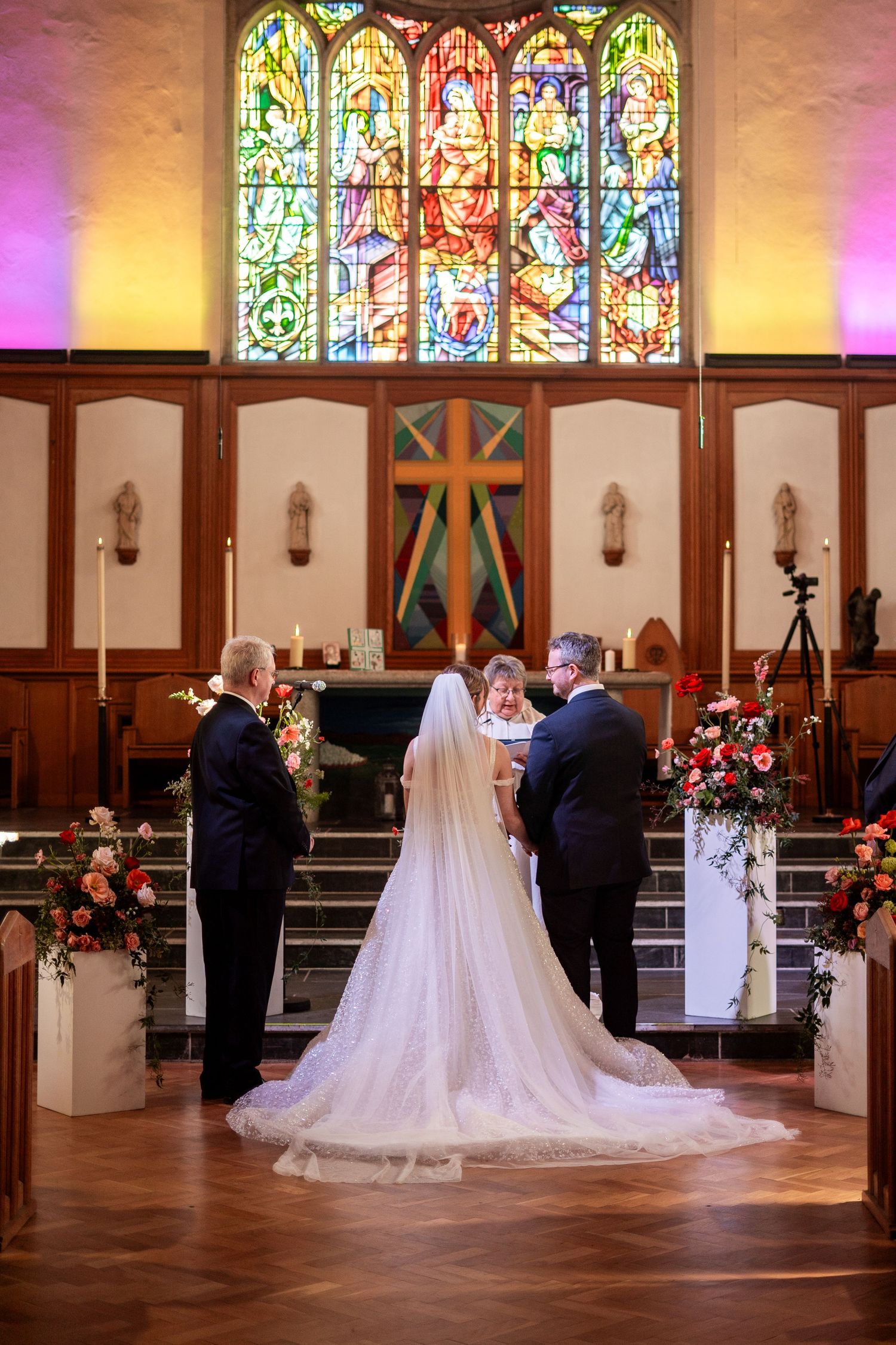 Bride and groom exchange vows at church altar in Penzance Church with stunning stained glass window and floral arrangements.