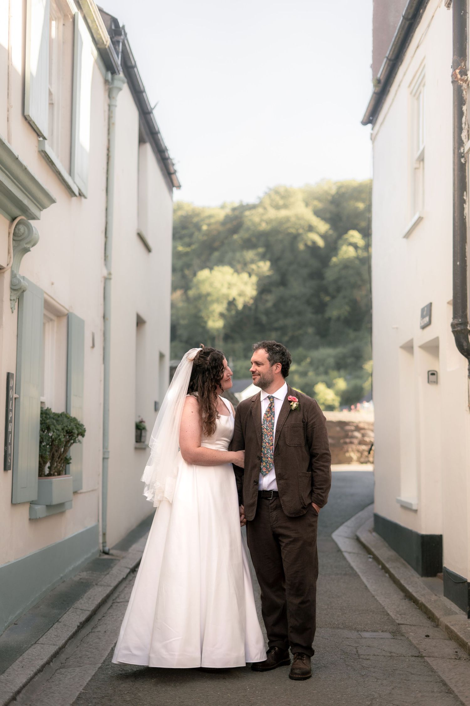 Bride and groom smiling at each other whilst walking through their local village in Cornwall