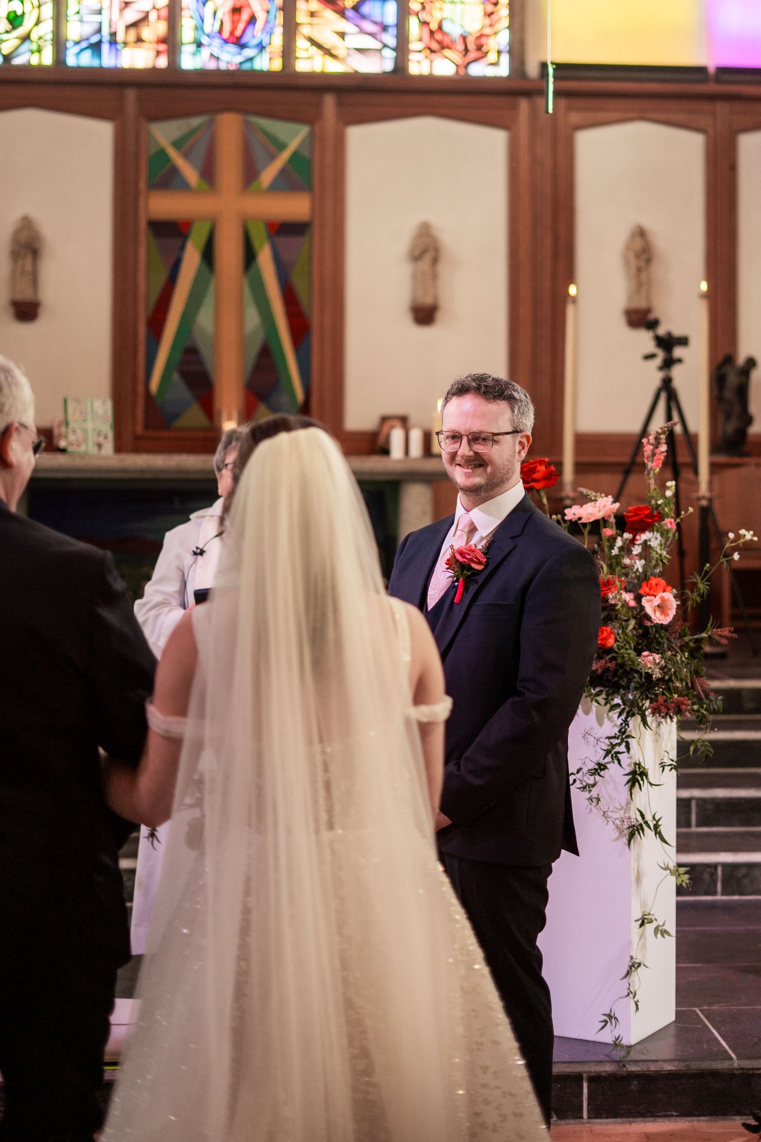 Groom watching the bride walk down the isle in Penzance Church amid stunning stained glass window and floral arrangements.