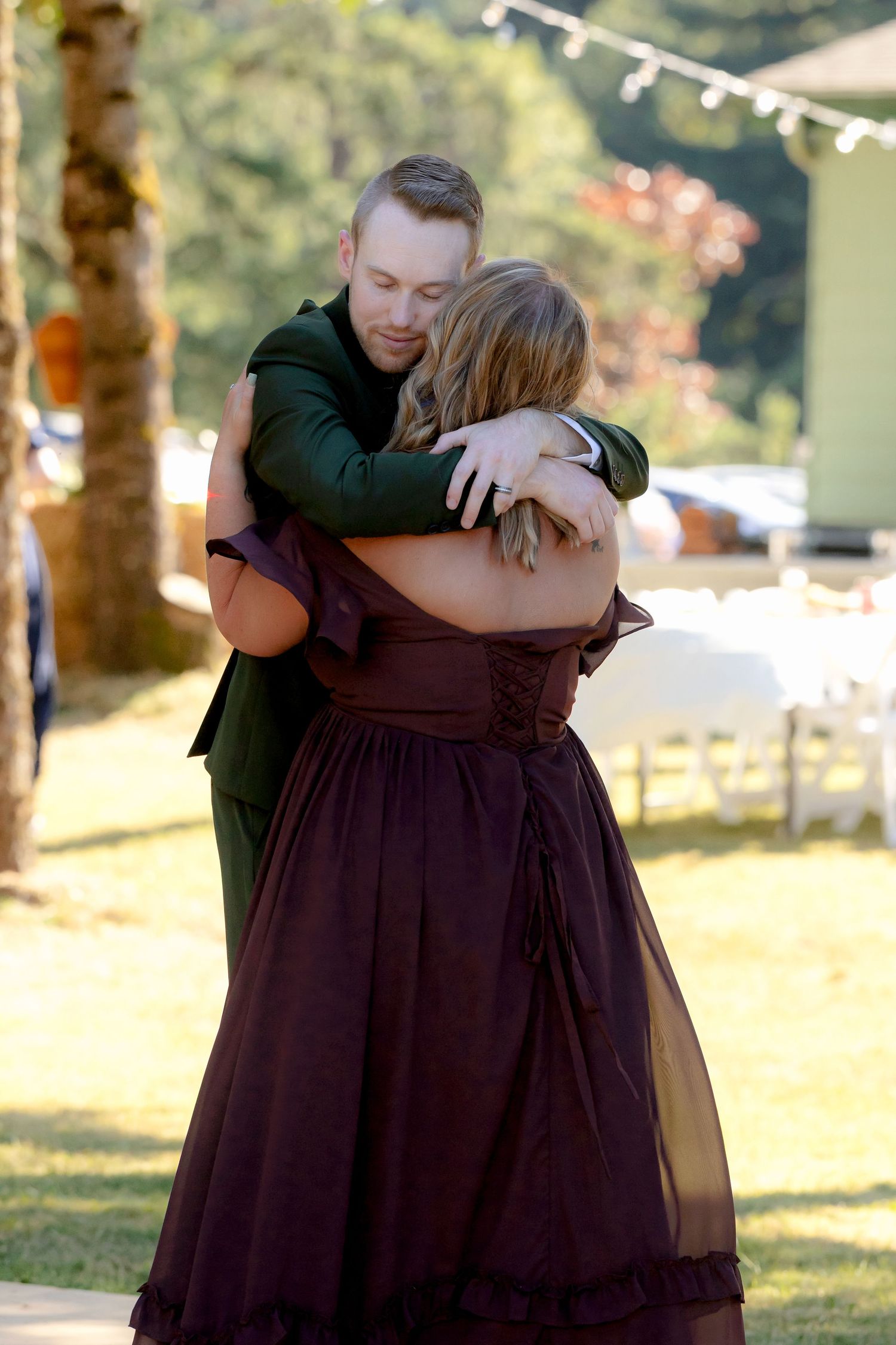 Groom in green suit shares emotional hug with woman in burgundy dress at outdoor wedding.
