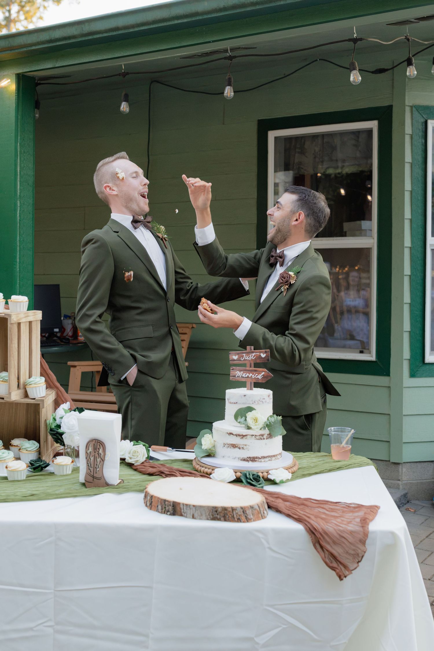 Two grooms in matching green suits share a playful cake feeding moment at their outdoor wedding reception.
