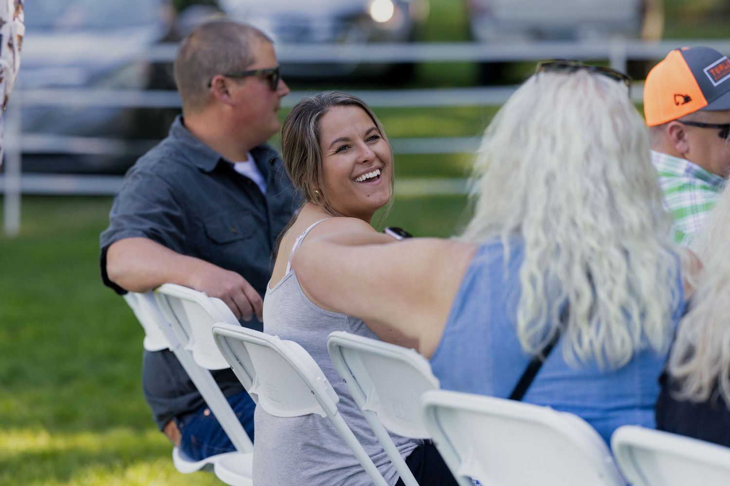 People laughing and socializing on white folding chairs at an outdoor event on a sunny day.