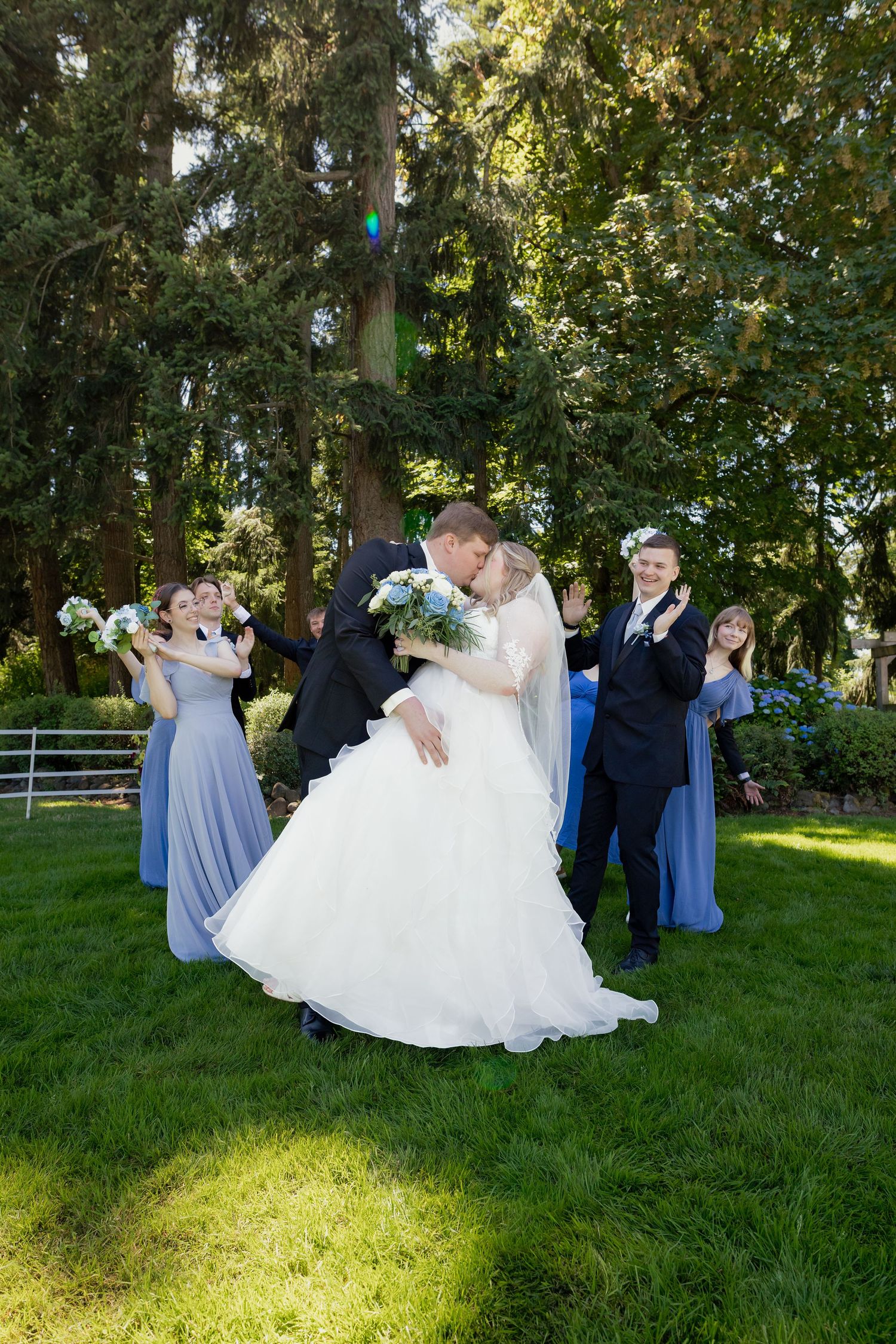 Newlyweds share a kiss surrounded by cheering bridesmaids in blue and groomsmen on a green lawn.