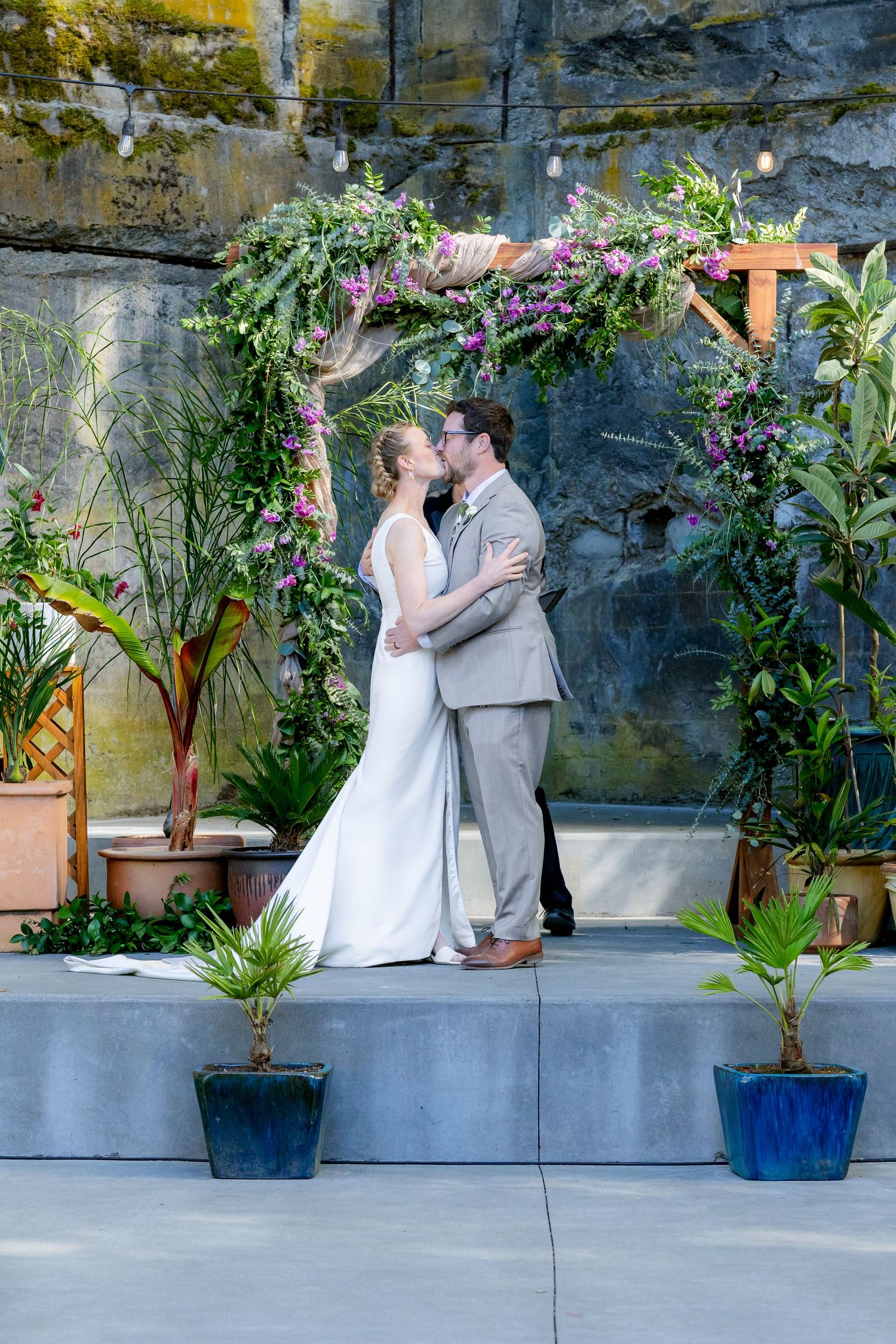 Bride and groom share first kiss under a floral arch adorned with purple flowers and greenery at outdoor wedding ceremony.