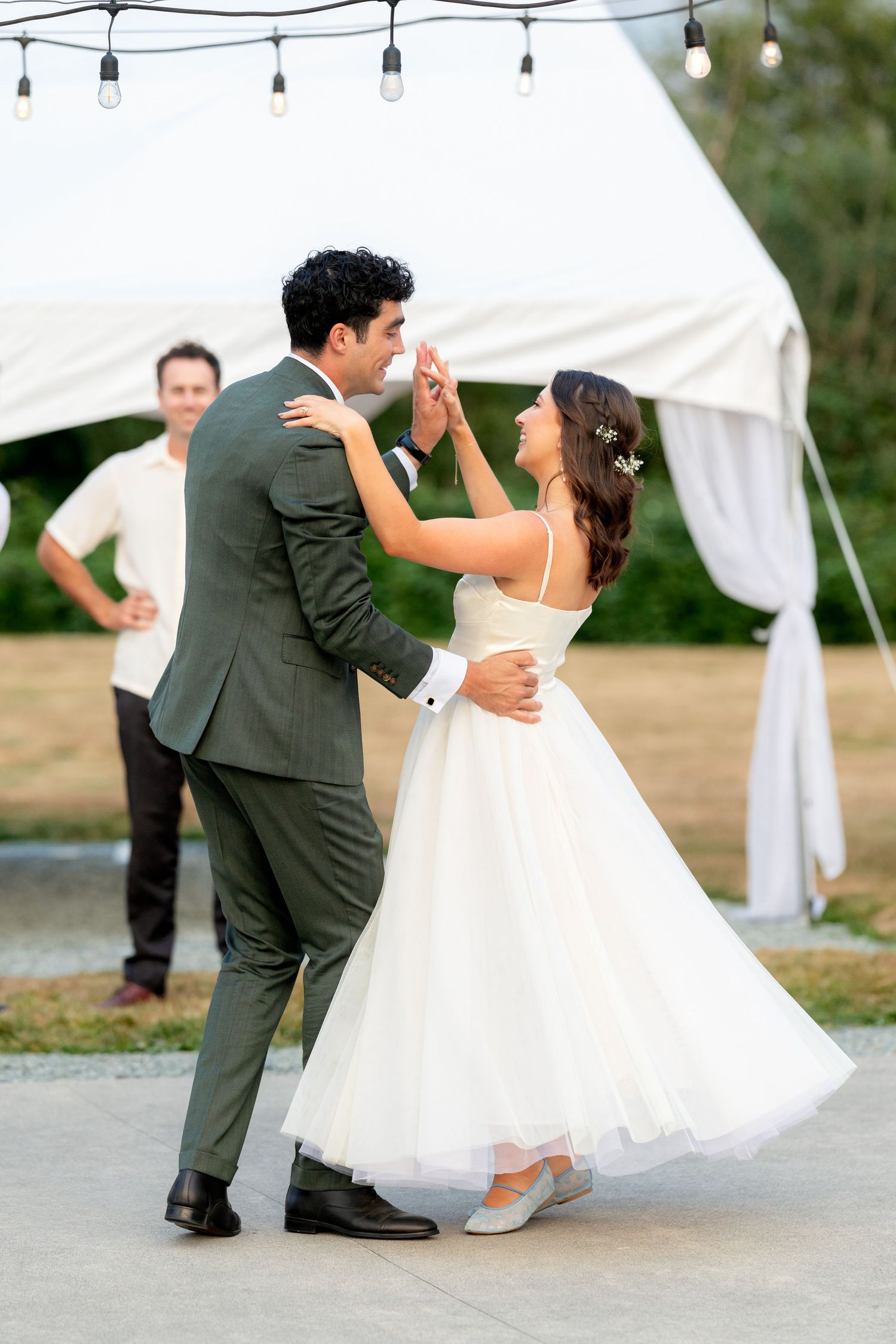 Bride and groom share first dance outdoors under string lights near white tent, bride in tea-length gown, groom in green suit.