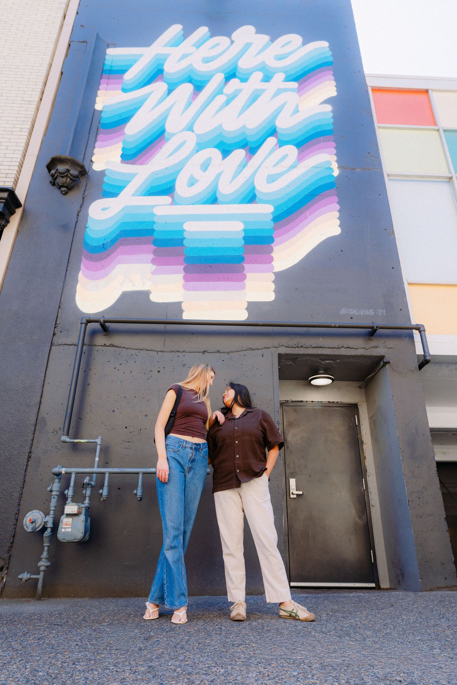 Two people stand beneath a colorful Here With Love mural on a building exterior, sharing a romantic moment.