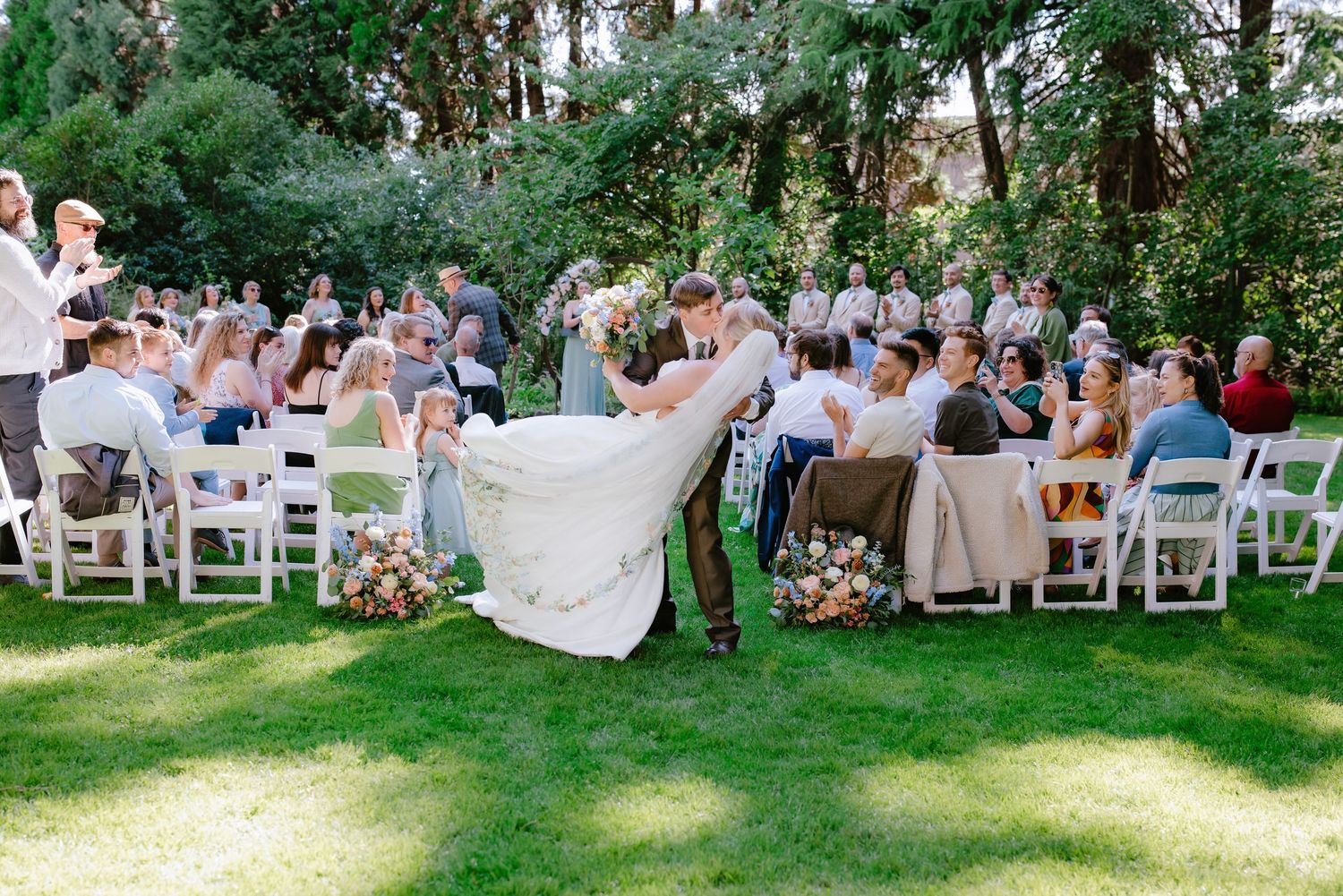 Groom dips bride for a kiss at outdoor wedding ceremony on green lawn surrounded by seated guests.