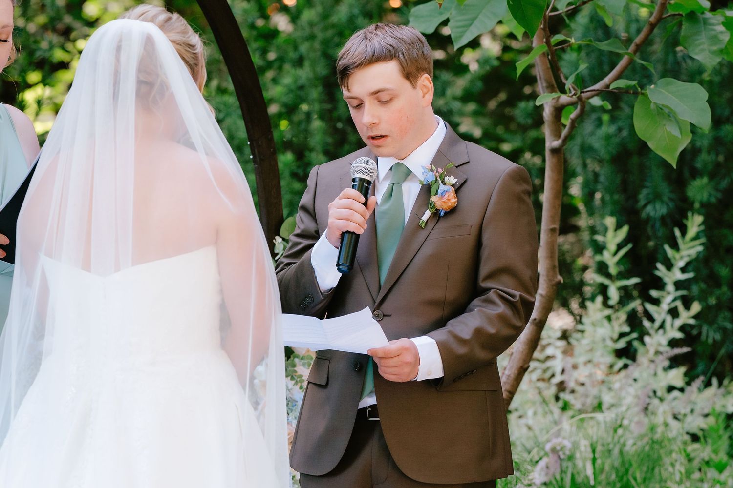 Groom reads vows from paper at outdoor wedding ceremony, wearing brown suit with green tie and boutonniere.