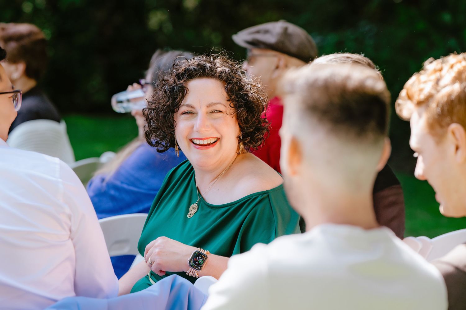 Woman in a green off-shoulder dress laughing joyfully while seated at an outdoor wedding ceremony.
