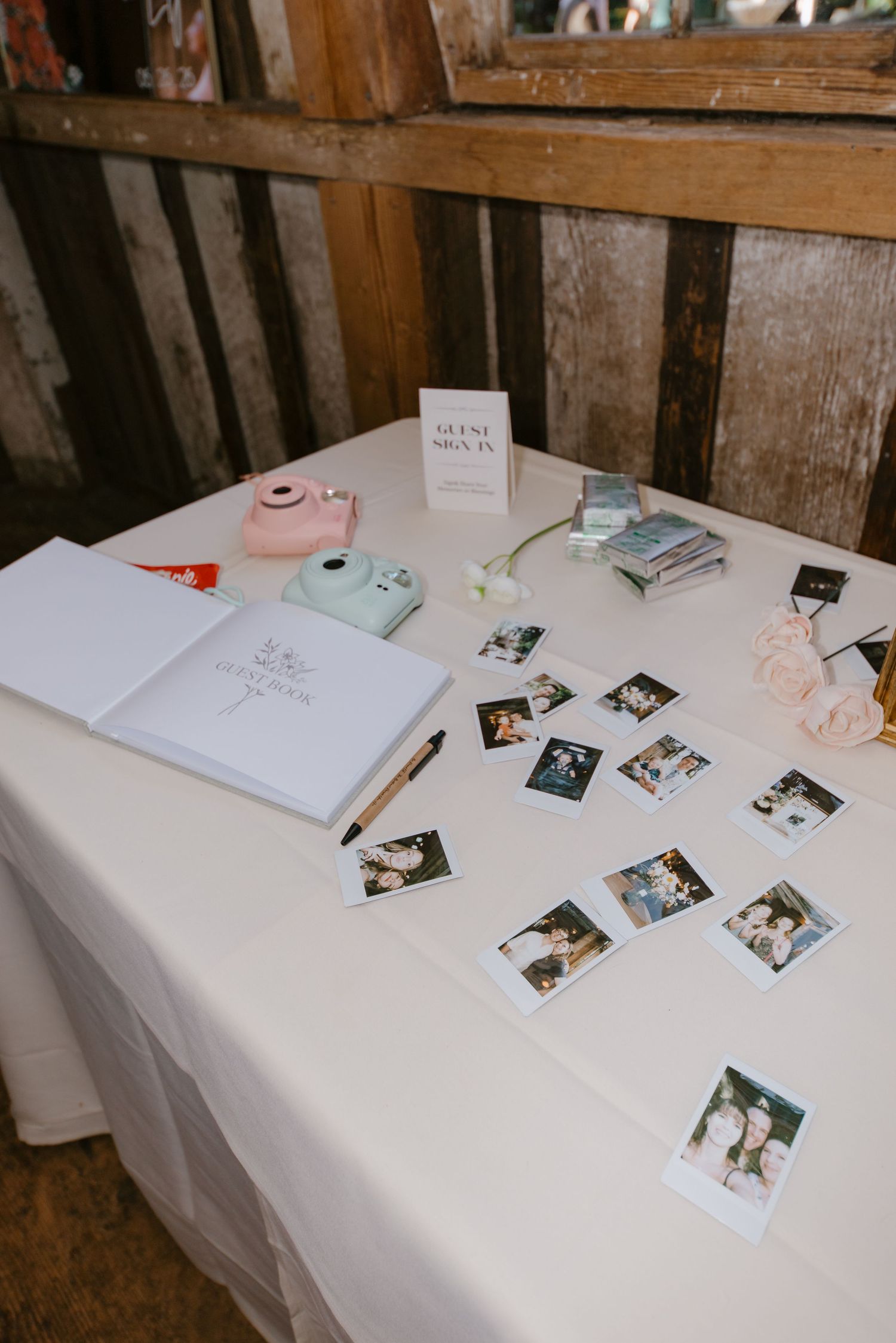 Wedding guest book table with Polaroid photos, Instax camera, white guestbook, and floral decor on white linen.