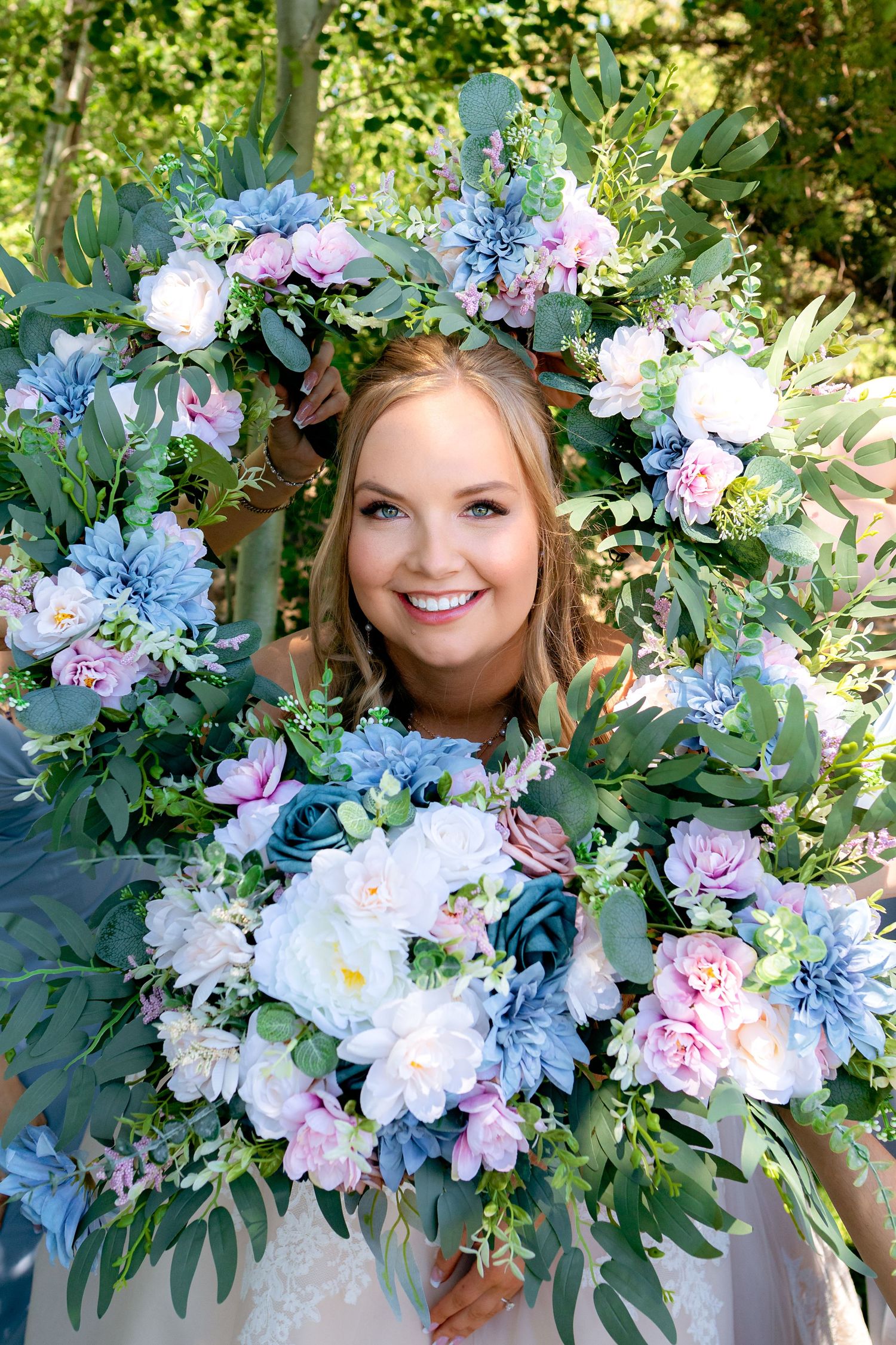 Smiling bride framed by lush blue pink white floral arrangement holding bouquet outdoors.