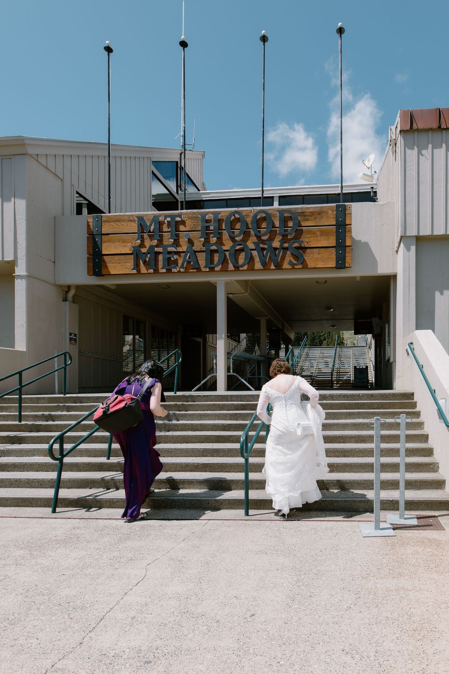 Bride in white gown and guest climbing stairs at Mt. Hood Meadows ski resort entrance on a sunny day.