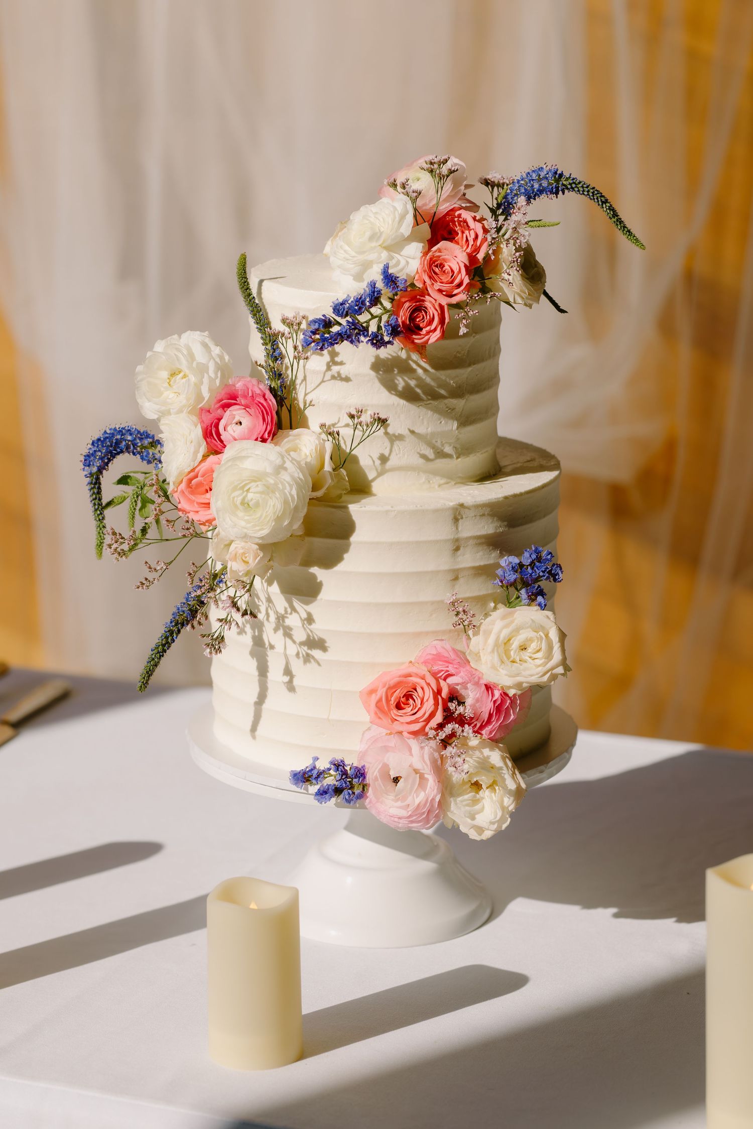 Three-tier white textured wedding cake adorned with pink, white, and blue fresh flowers.