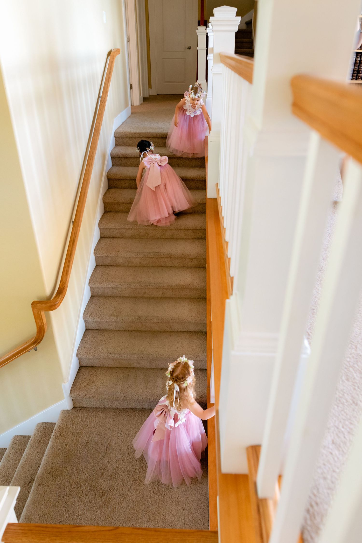 Three flower girls in pink tulle dresses climbing carpeted stairs with white banister at a wedding.