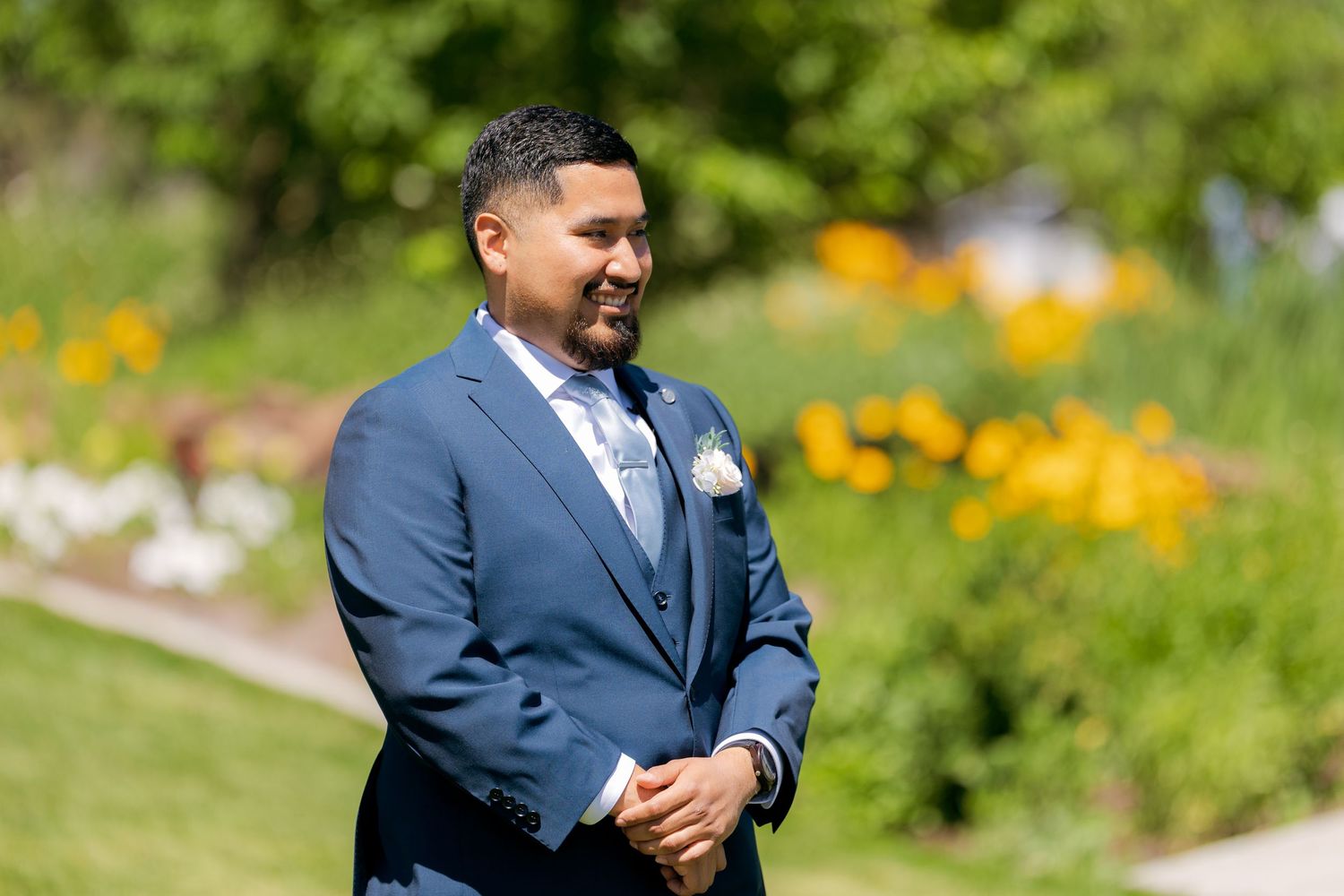Smiling groom in blue suit with boutonniere stands outdoors with colorful flower garden in background.