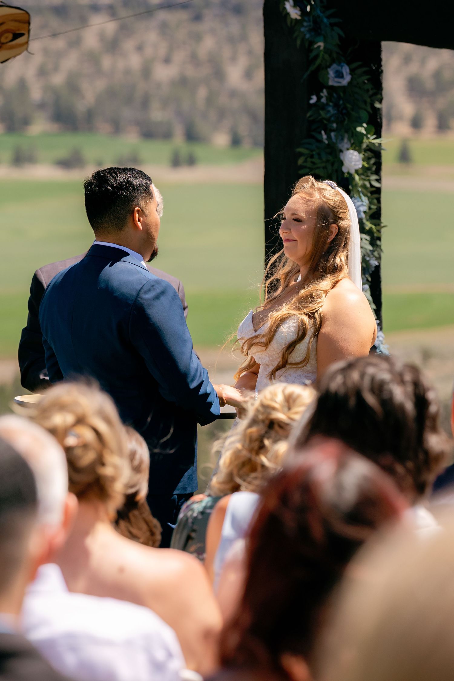 Bride and groom exchange vows at outdoor wedding ceremony with scenic green valley backdrop.