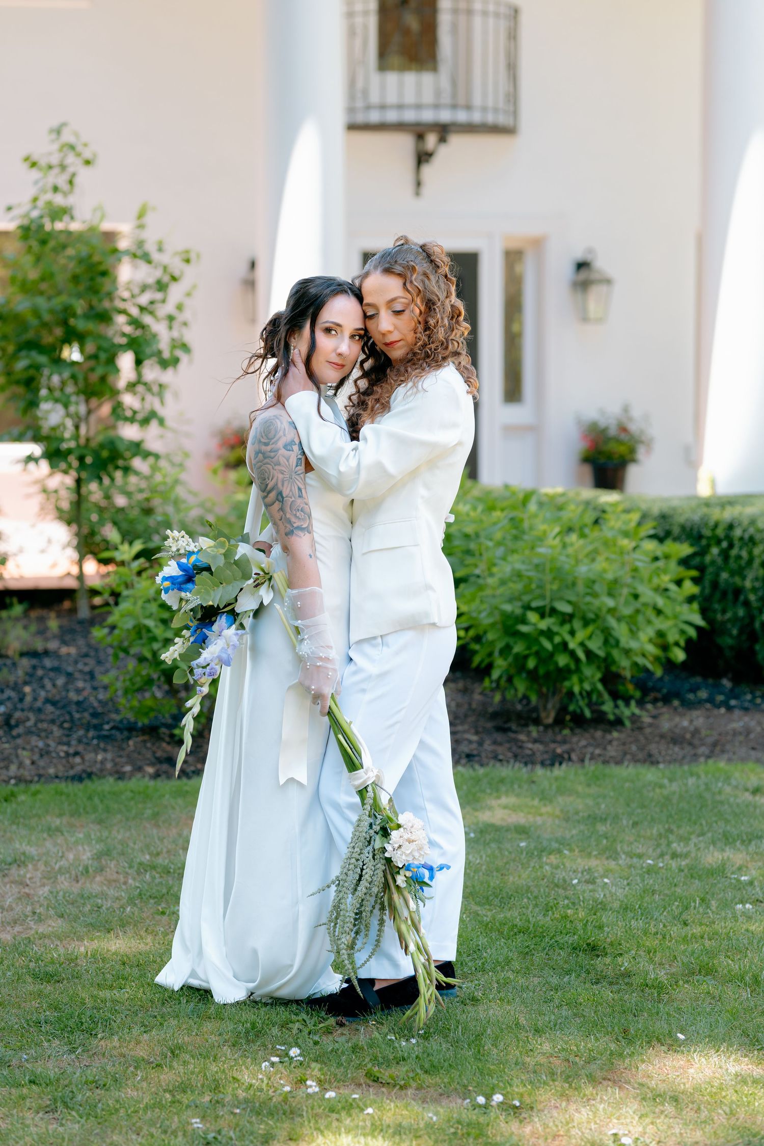 Two brides in white wedding attire embrace outdoors, one in a gown with tattoos, holding blue and white flowers.