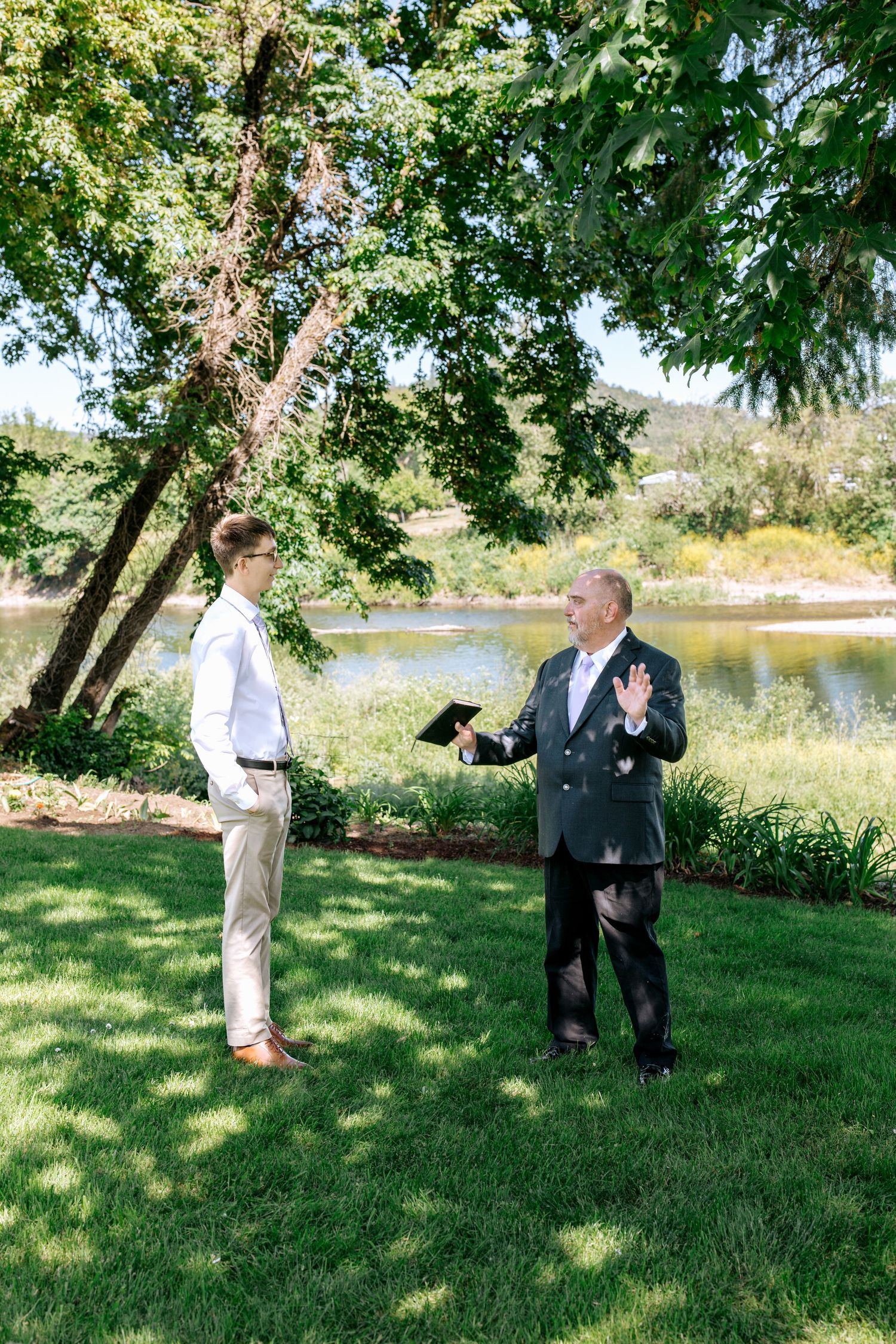 Officiant gestures while speaking with a young man in a white shirt near a scenic lakeside wedding venue.