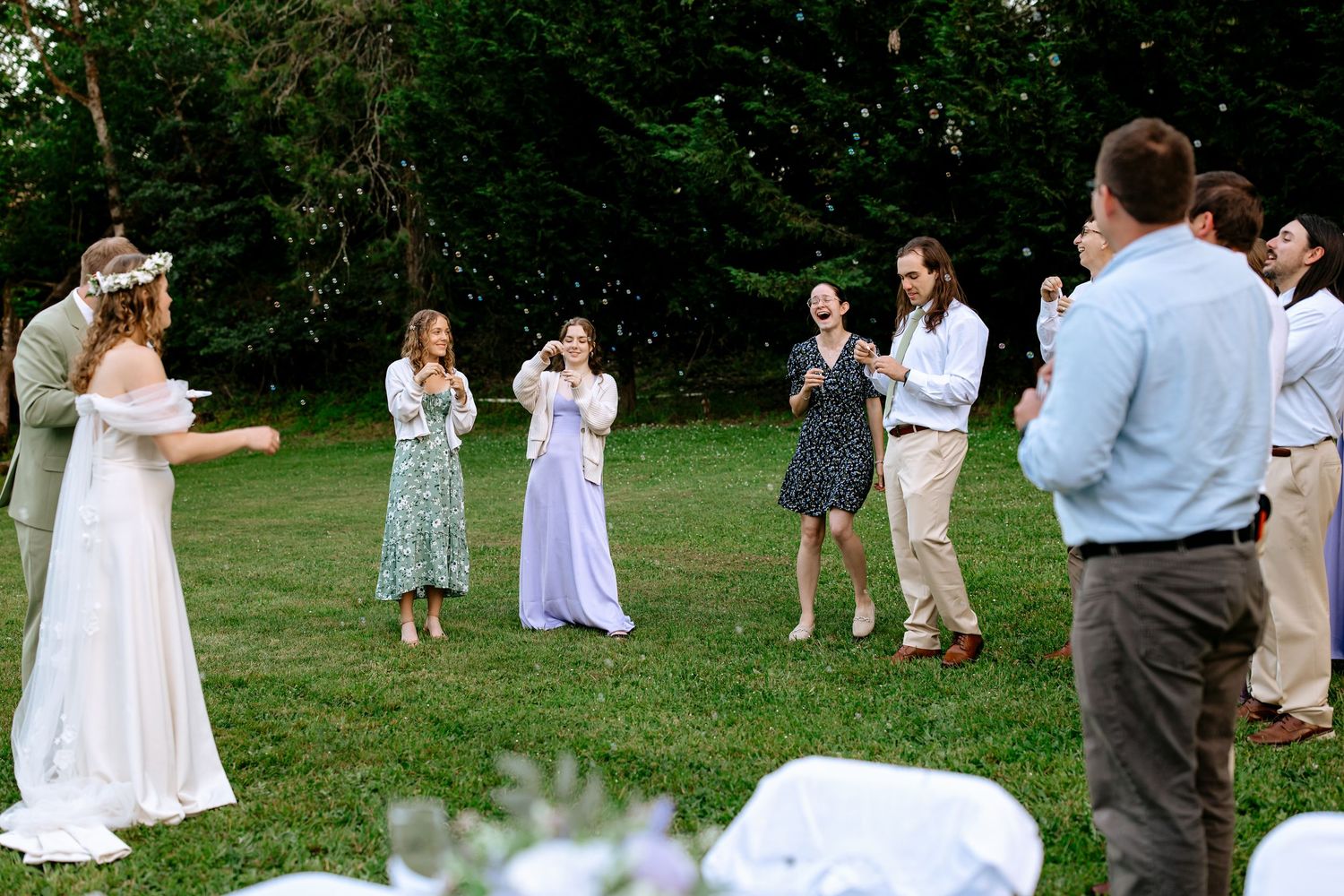 Bride in floral crown playing outdoor wedding reception game with guests on a sunny green lawn.