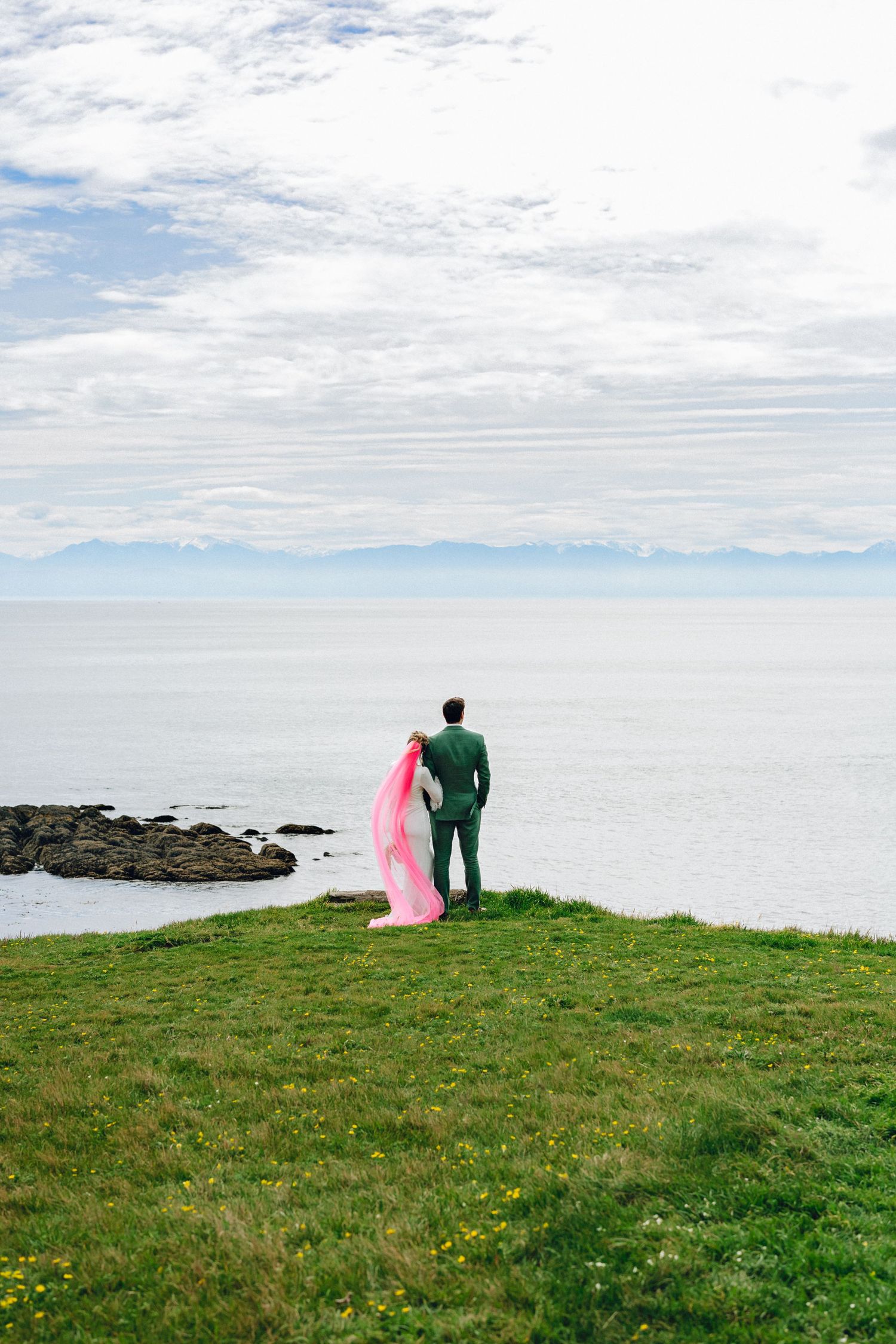 Groom in green suit stands on coastal cliff holding pink fabric, overlooking calm ocean with mountains in distance.