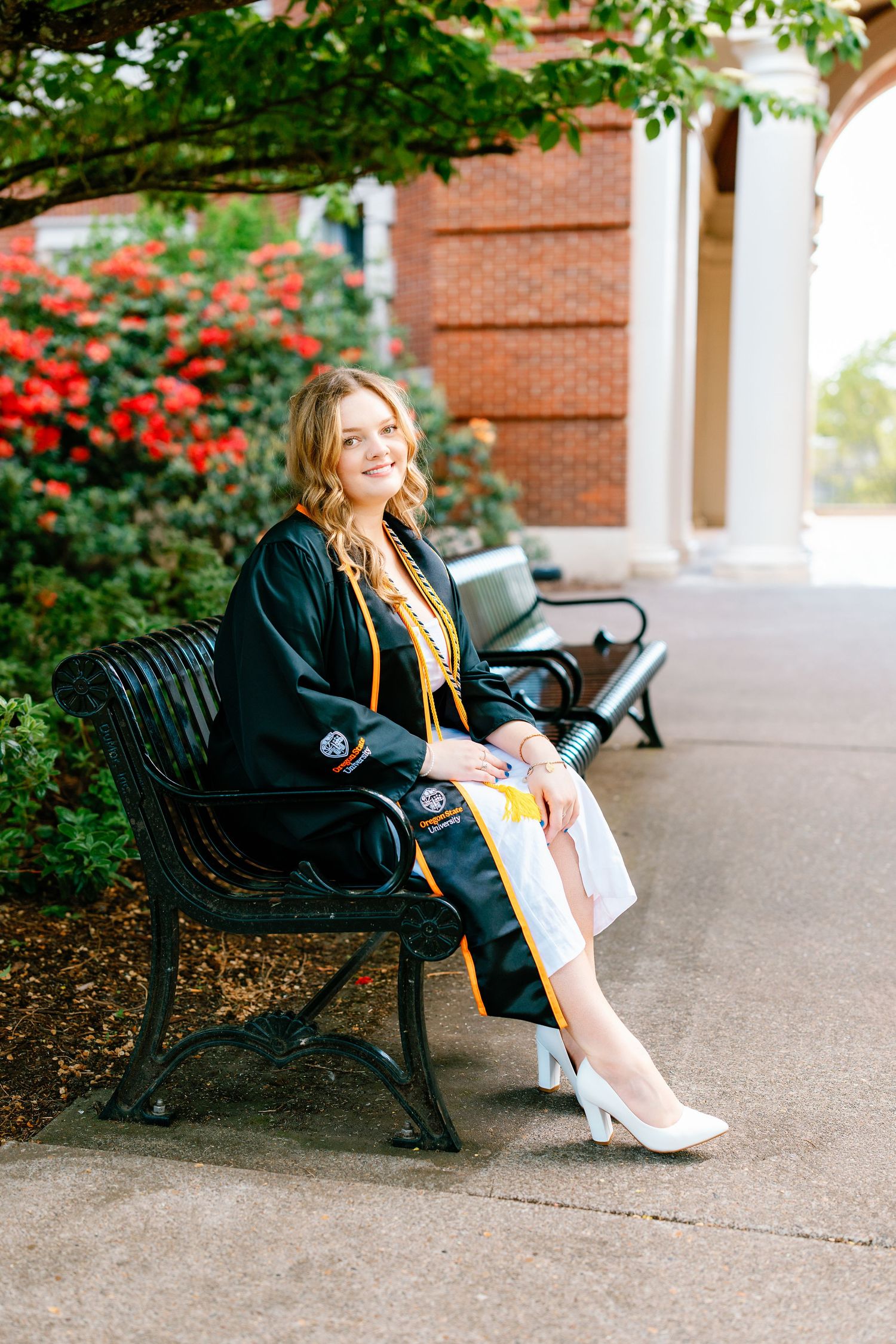 A smiling young woman in black graduation gown with gold accents sits on a bench outdoors near red flowers.