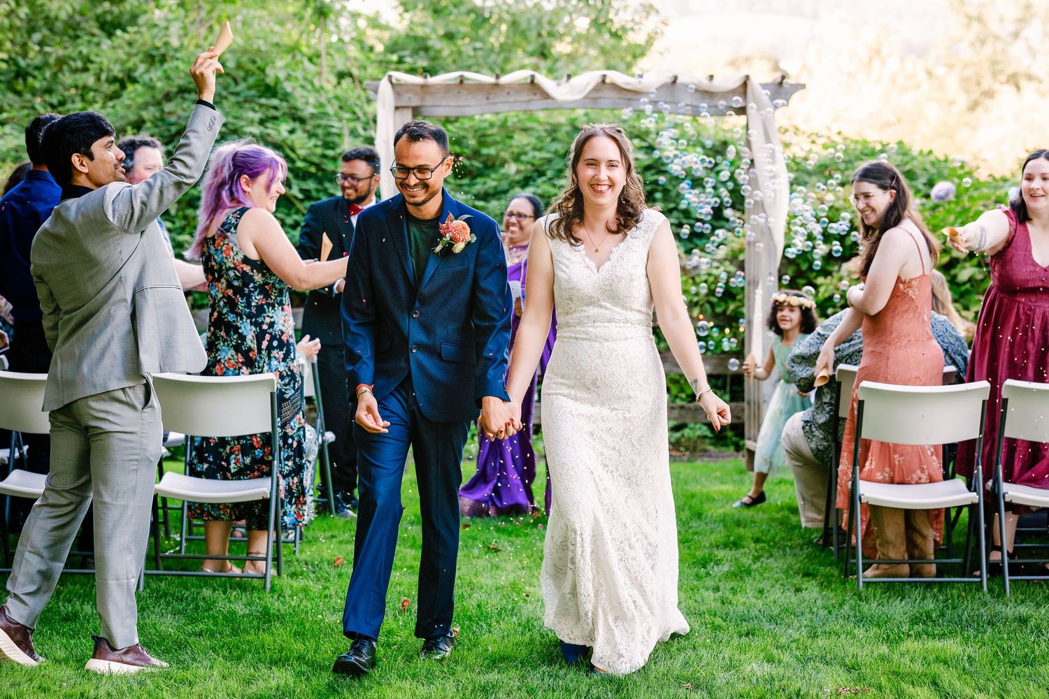 Newlyweds celebrate with guests during outdoor garden wedding ceremony with raised arms and joyful expressions.