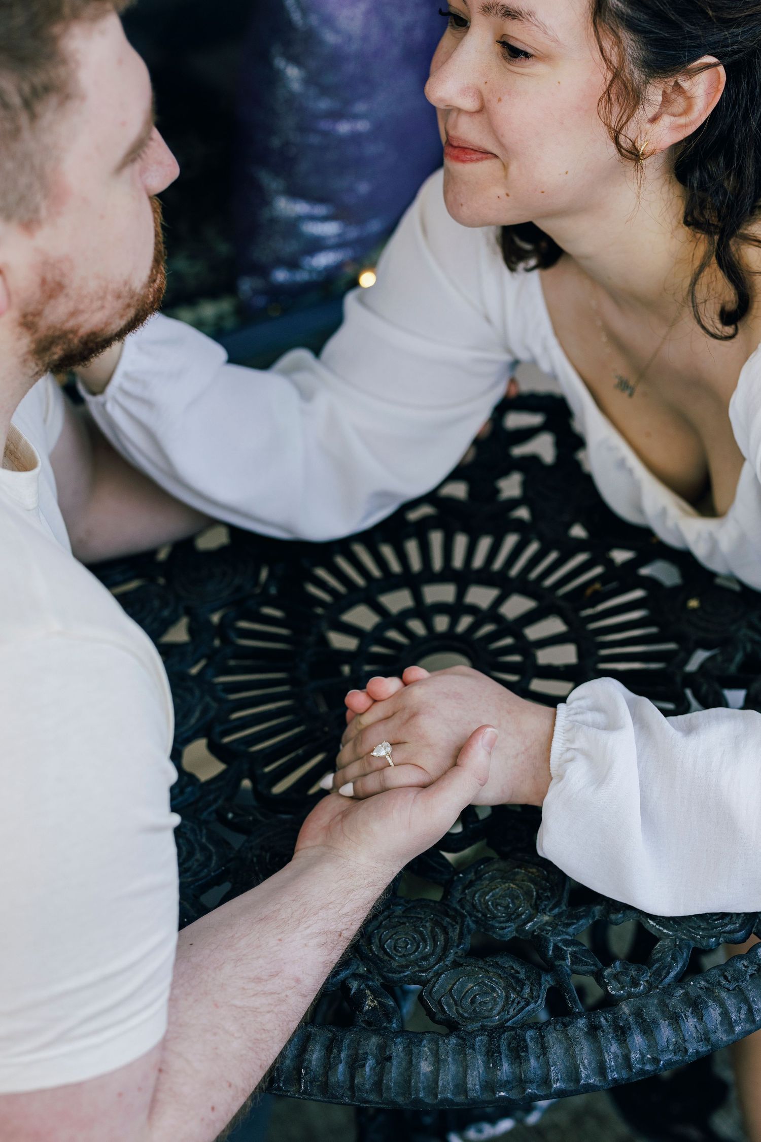 A couple in white clothing holds hands intimately across a decorative black metal table.