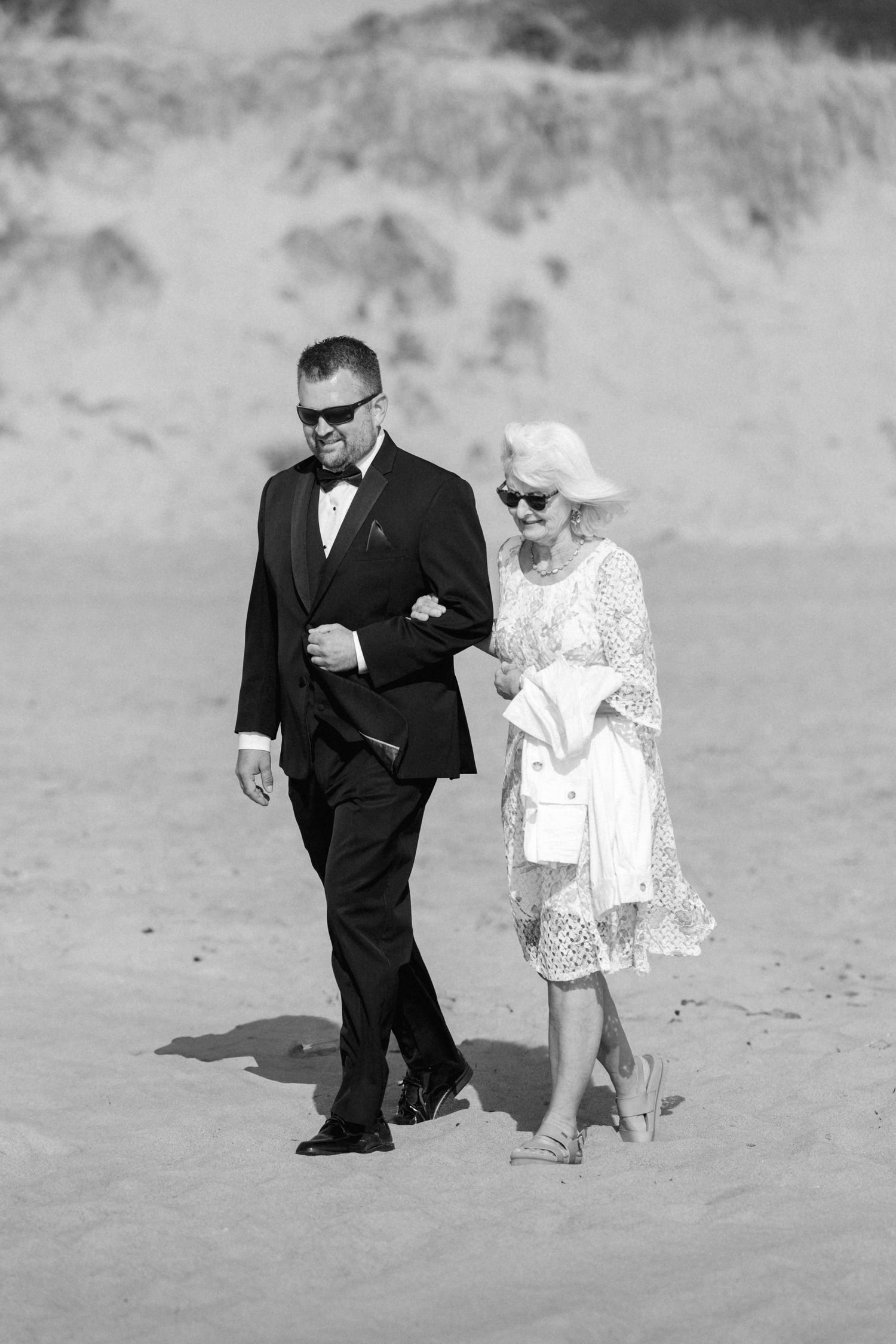 A black and white photo of a couple walking together on a beach in formal attire.