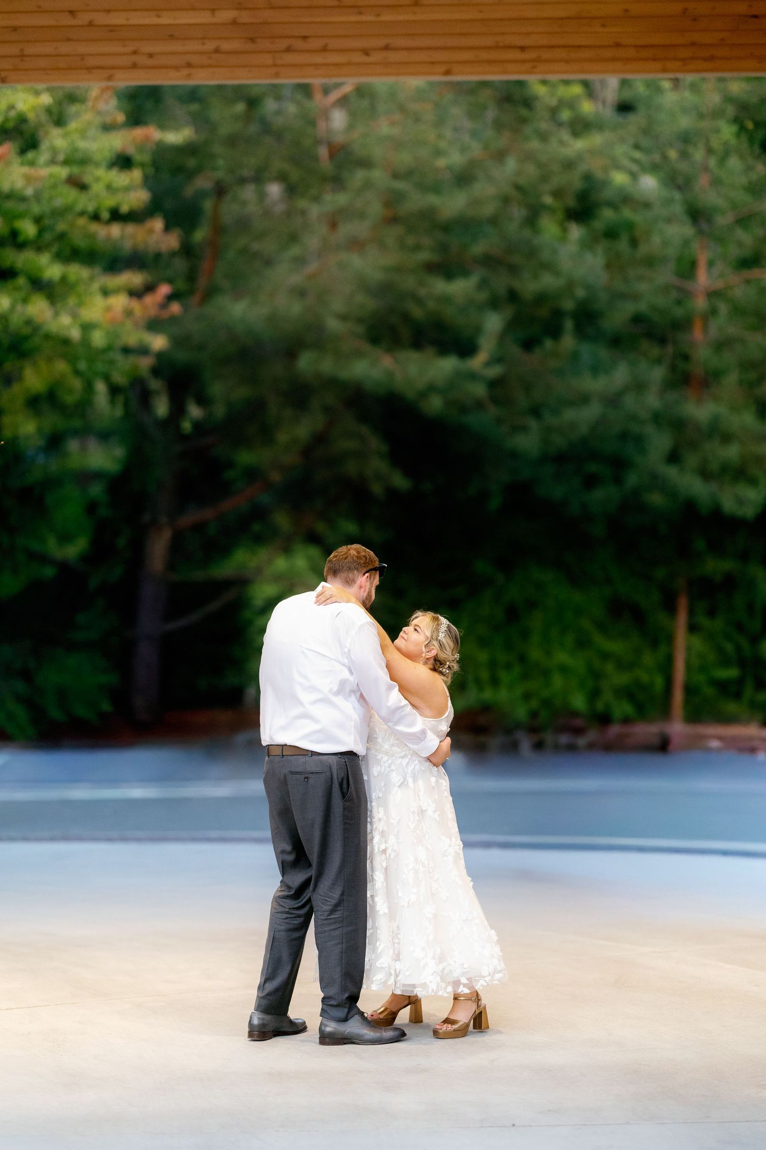 Wedding couple dances together in an outdoor parking area surrounded by green trees.